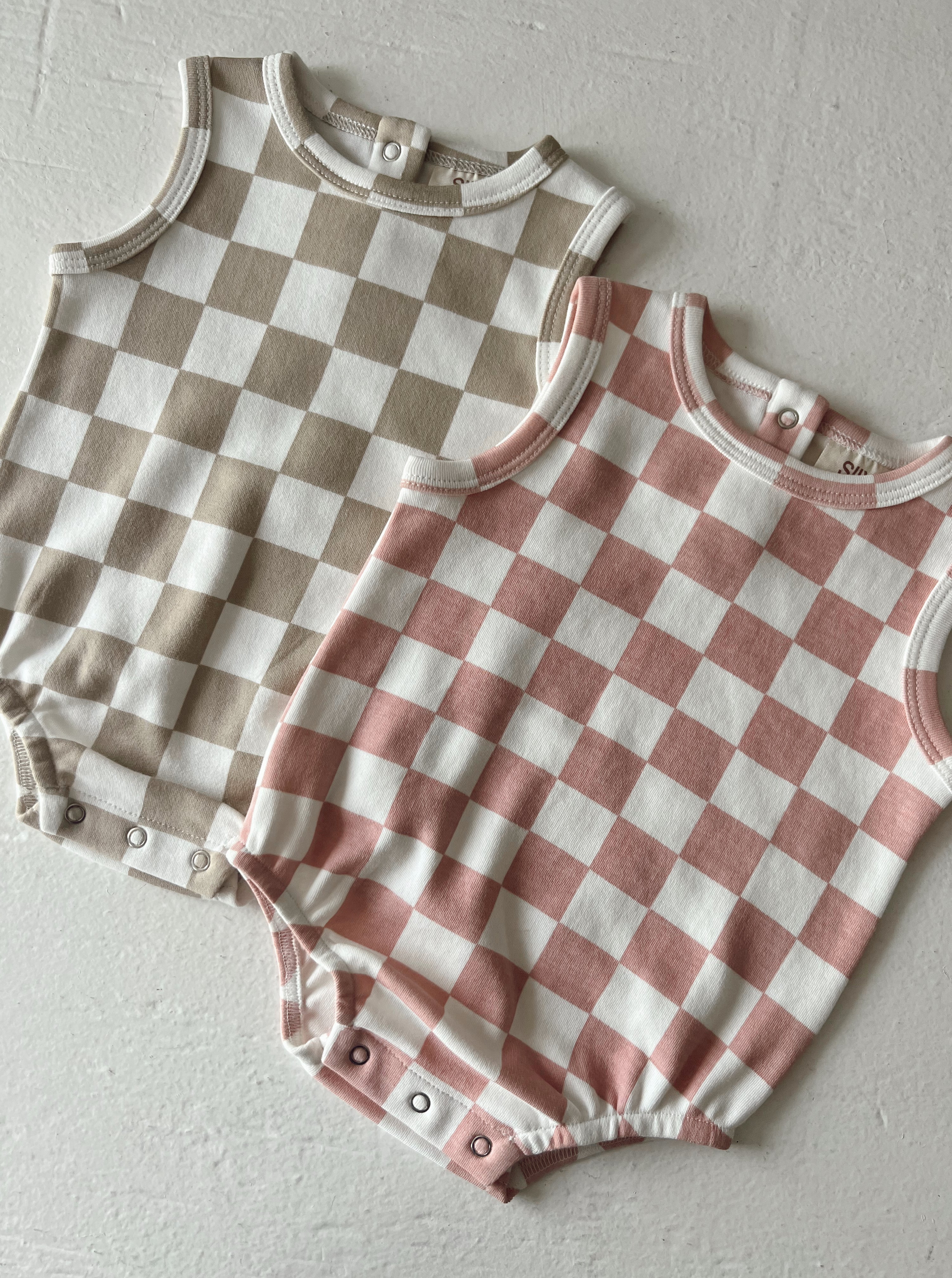 Two baby rompers with checkered patterns in beige and pink on a textured white backdrop.