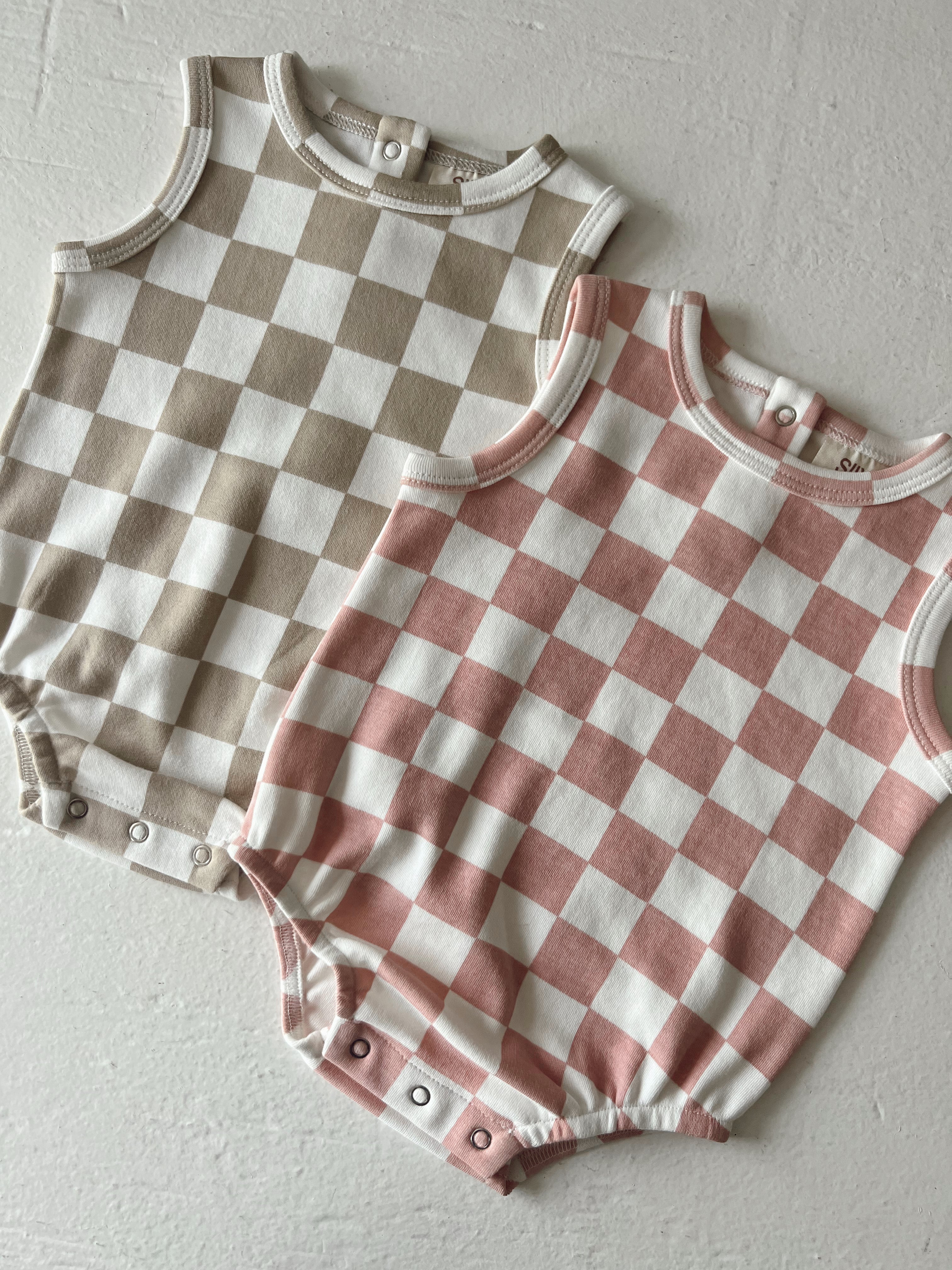 Two baby rompers with checkered patterns in beige and pink on a textured white backdrop.