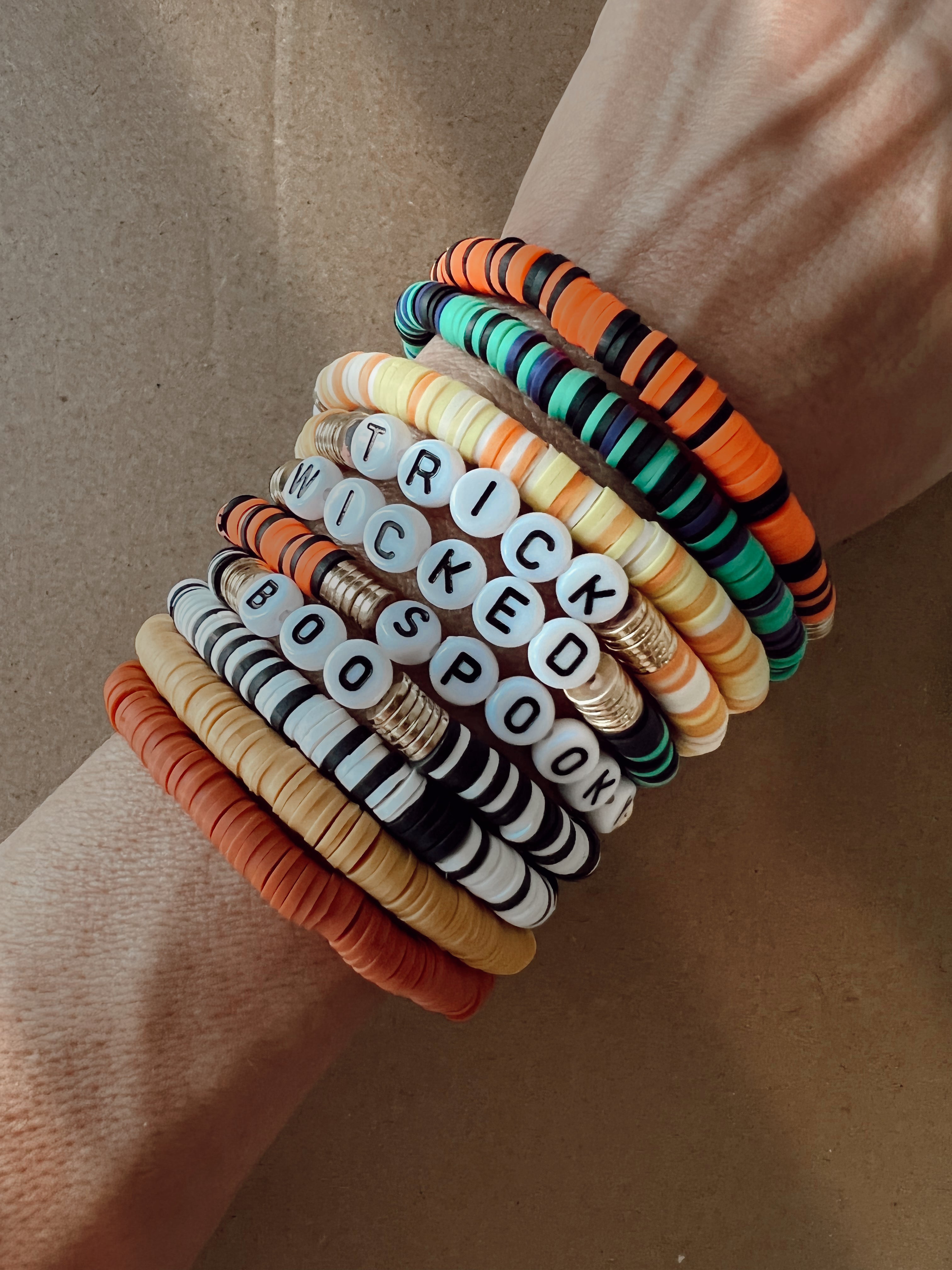 Colorful Halloween-themed bracelets stacked on a wrist, featuring words like "TRICK," "WICKED," "BOO," and "SPOOK."