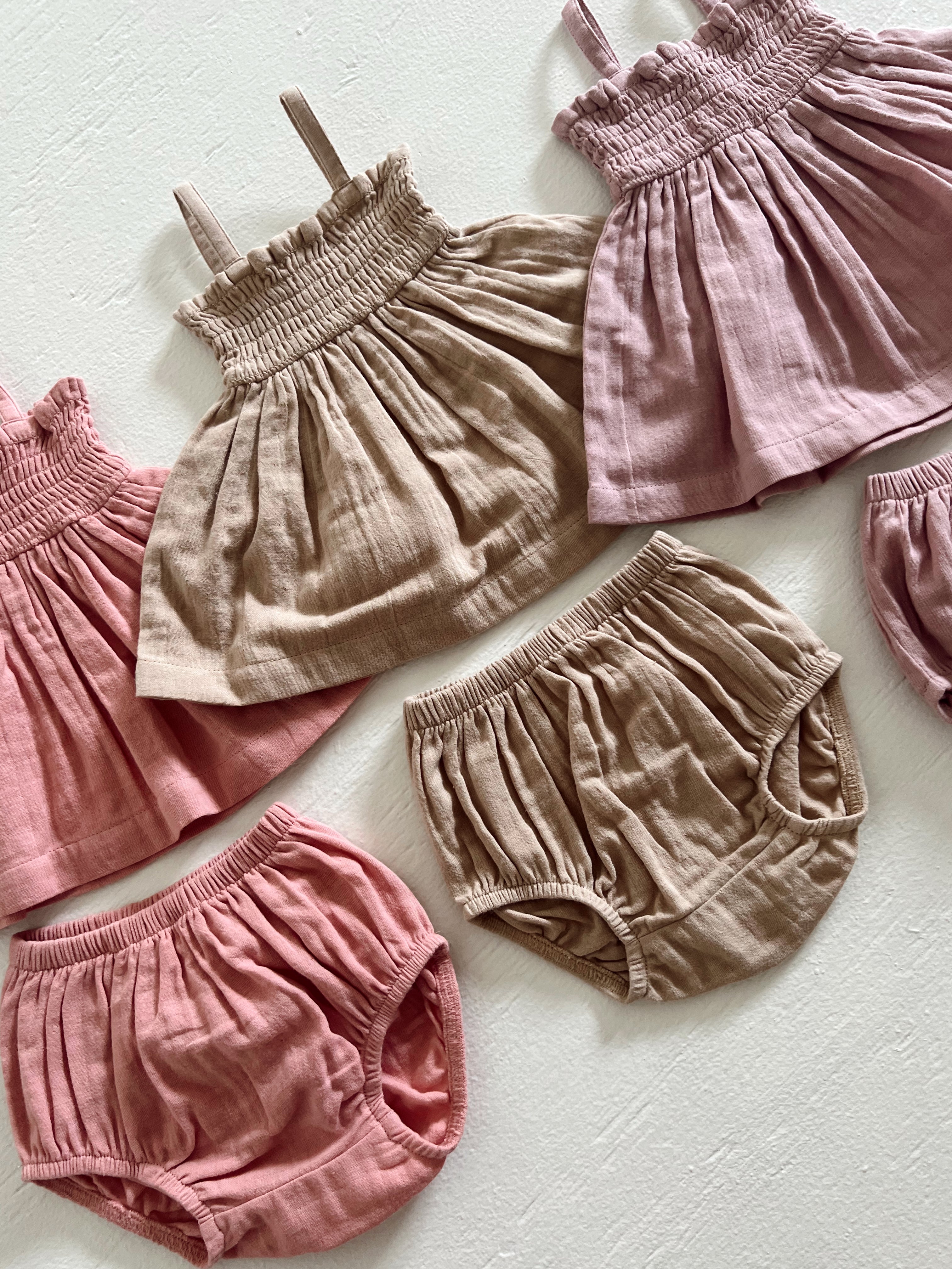 Assorted infants' linen tops and shorts in soft pink and beige hues on a light background.