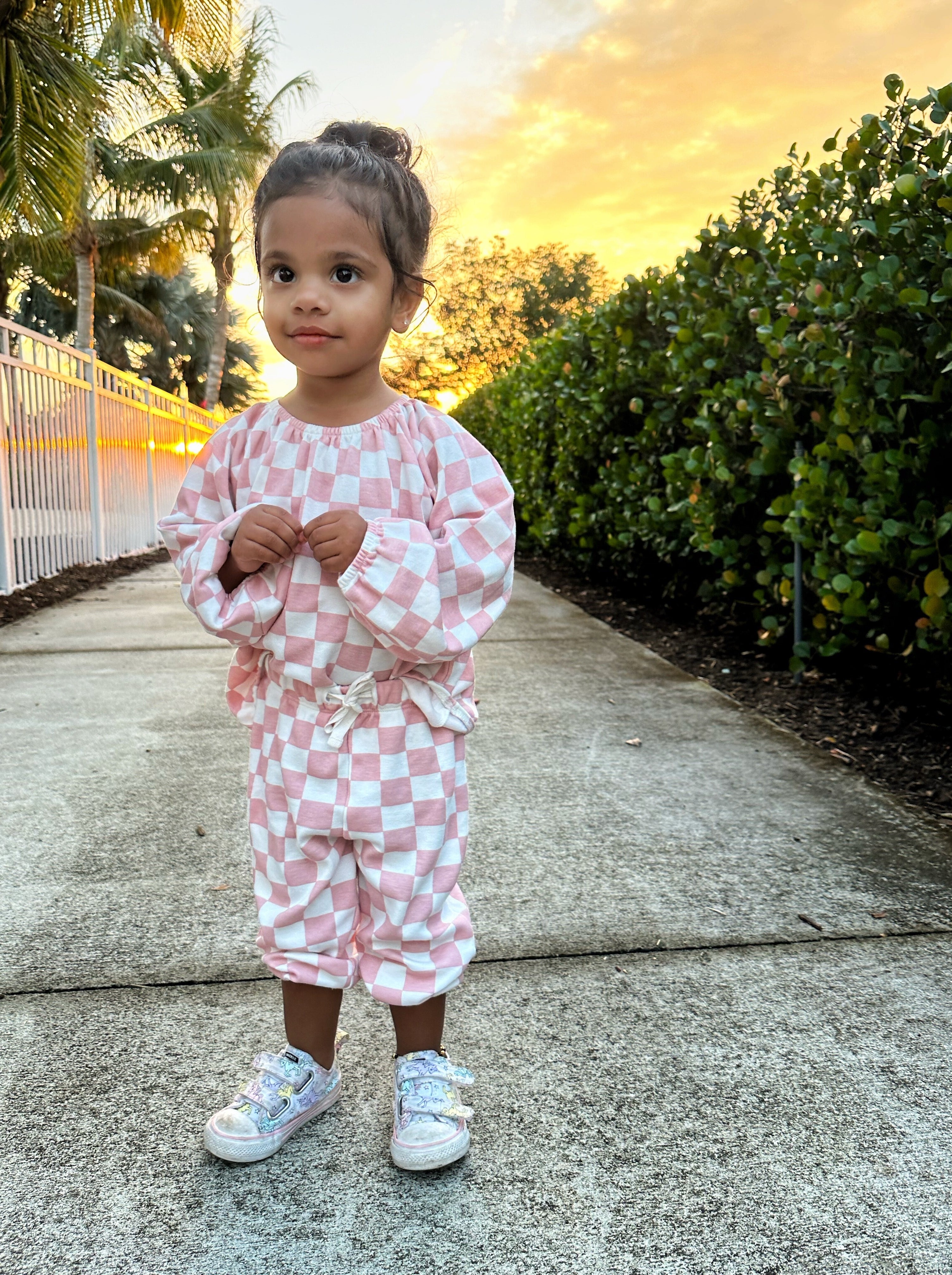 Child in pink checkered outfit stands on a sidewalk with a sunset backdrop and greenery.