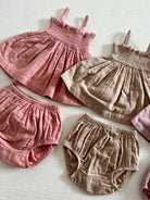 Collection of baby dresses and bloomers in pink and beige fabrics arranged on a light background.