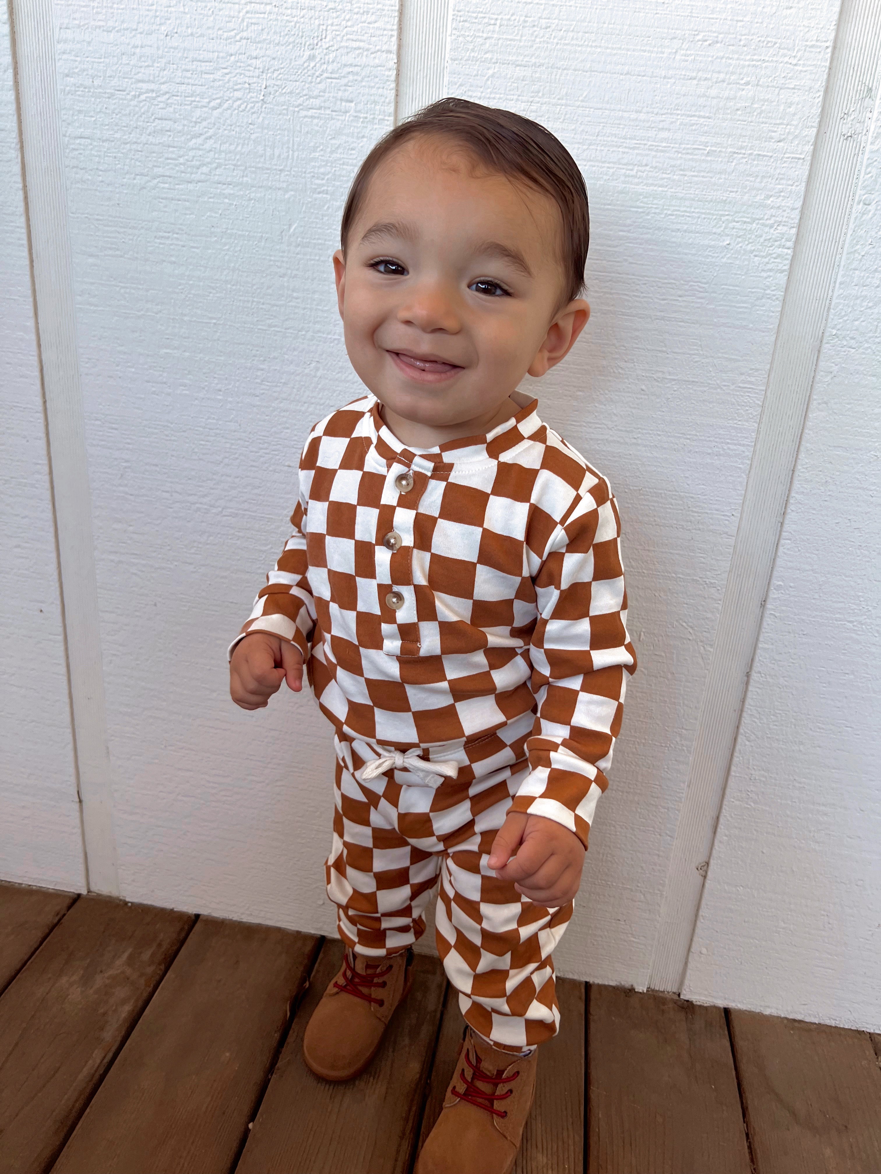 Smiling toddler in brown and white checkered outfit, standing on wooden floor with a white wall background.