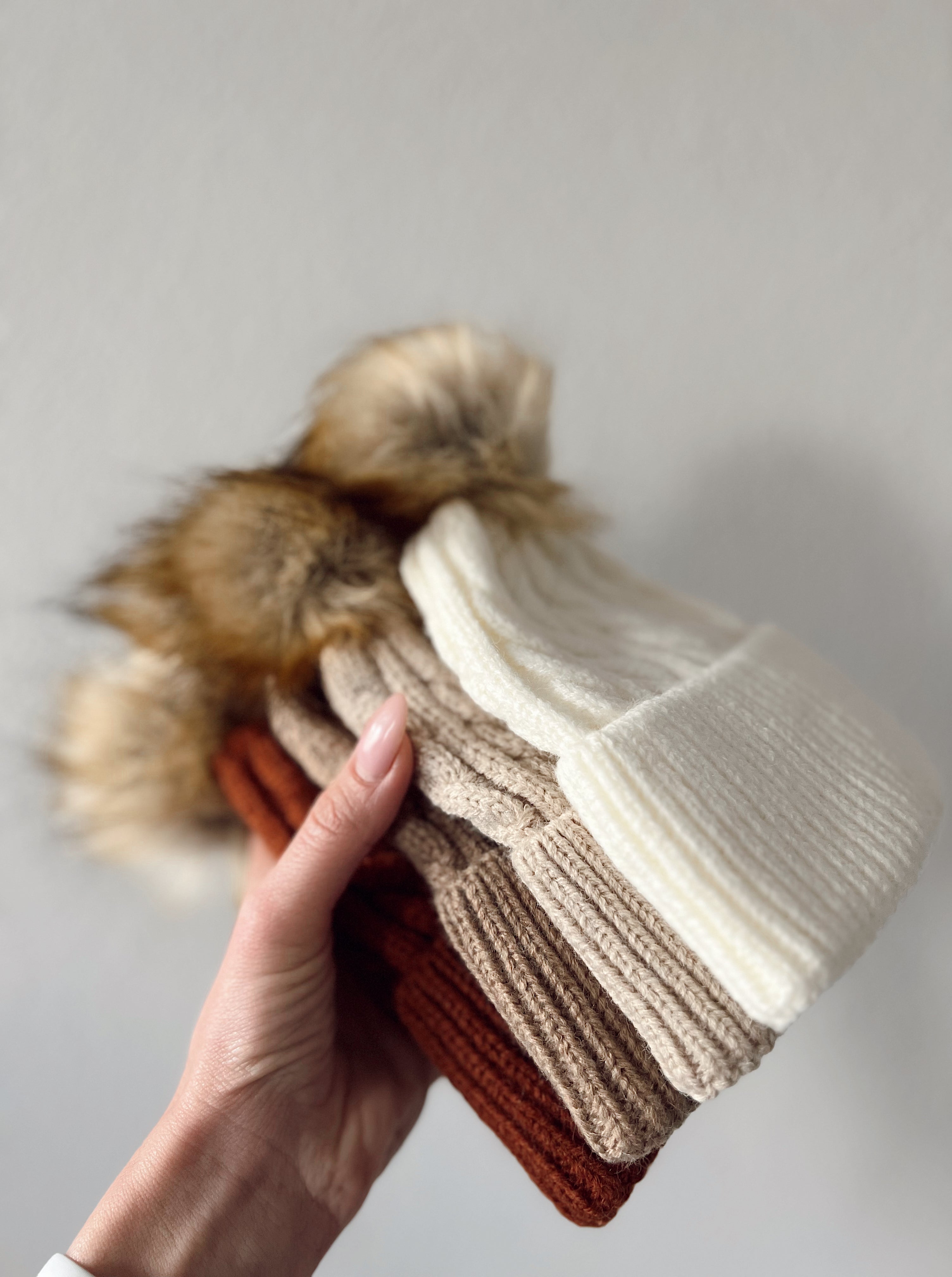 A hand holding four knit beanies in shades of cream, beige, brown, and rust with fur pom-poms.