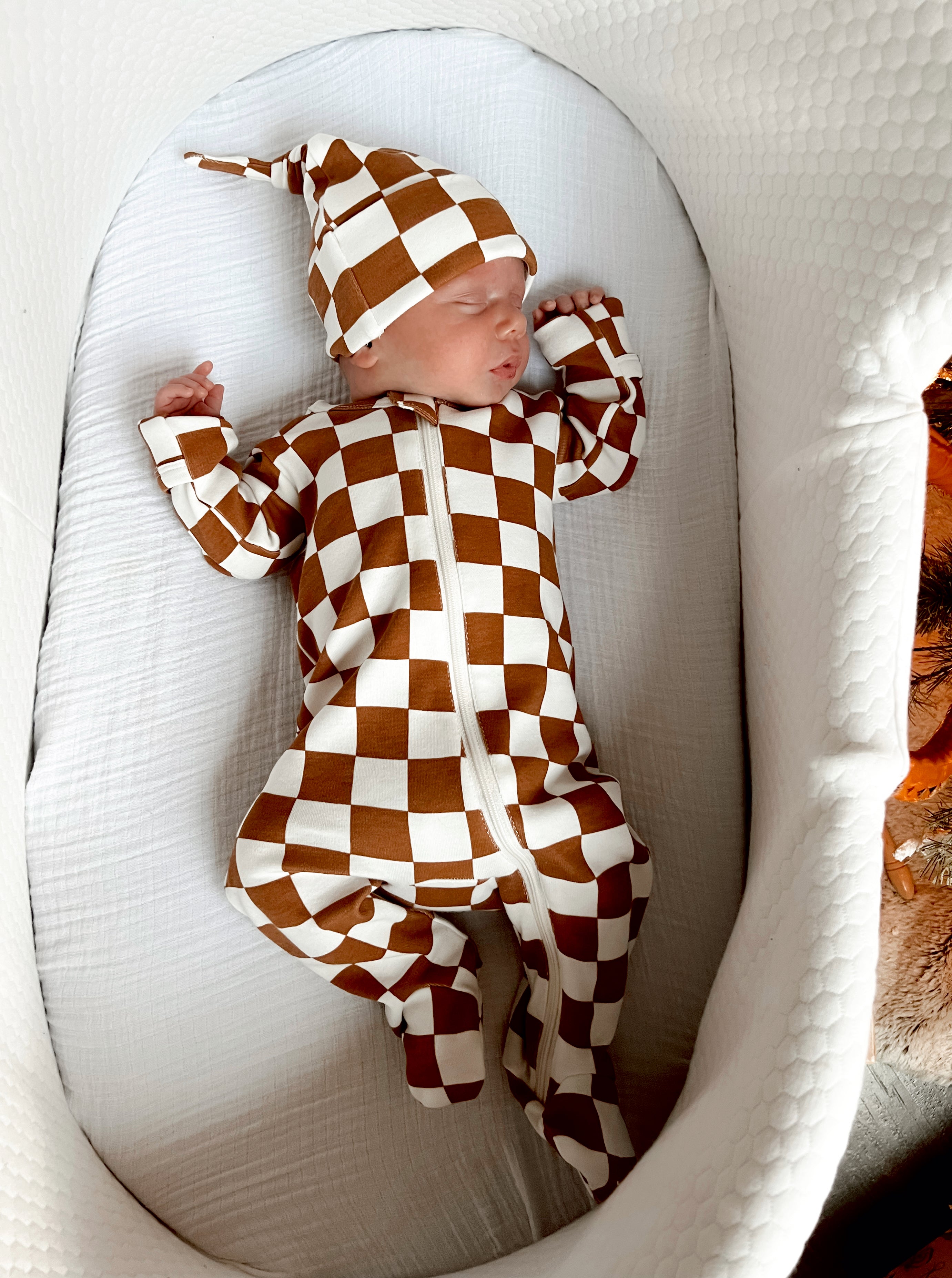 Infant sleeping in a brown and white checked onesie with a matching hat, nestled in a cozy bassinet.