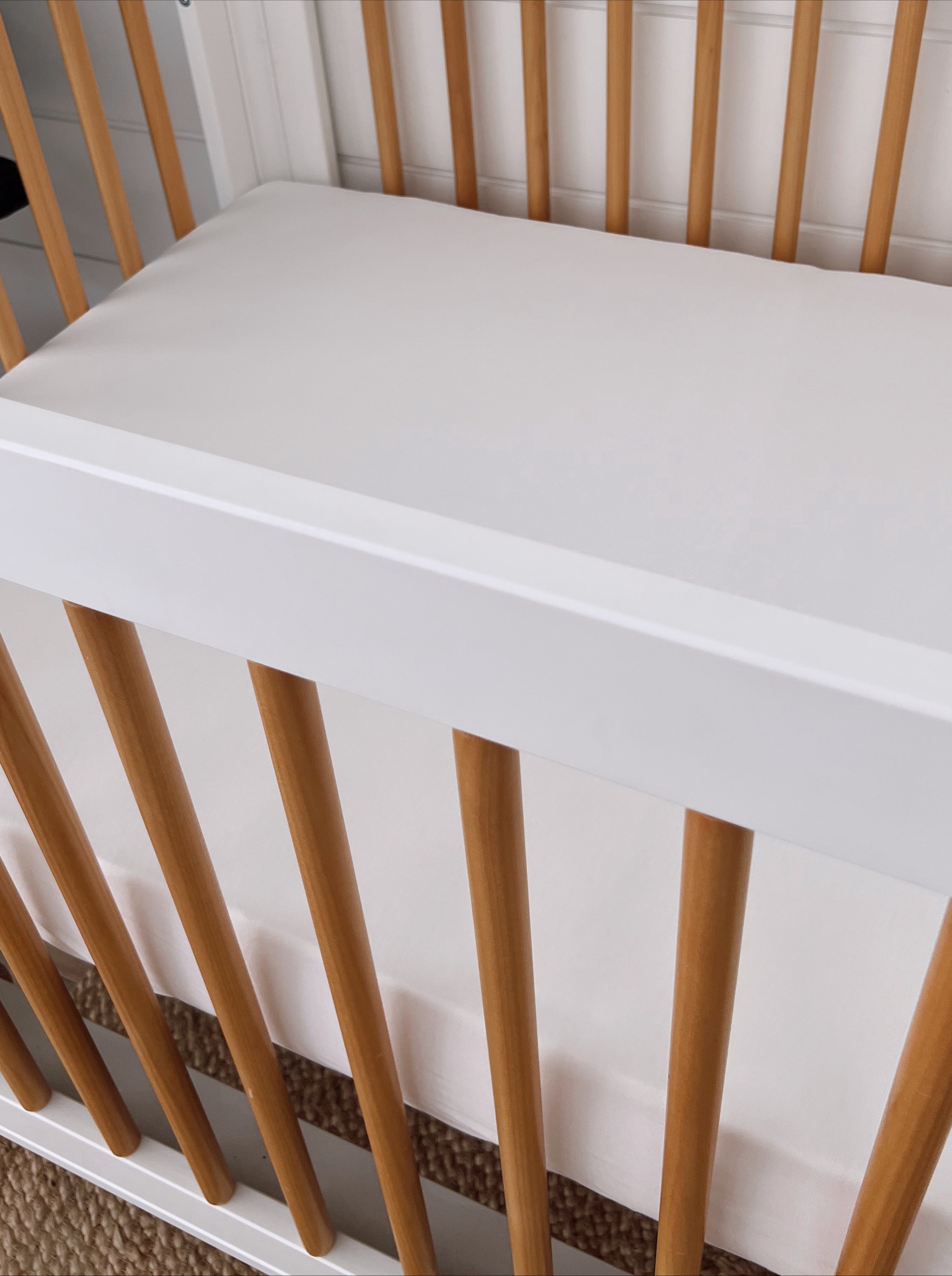 White crib with wooden slats and a plain mattress, set against a light background. Ideal for a nursery.