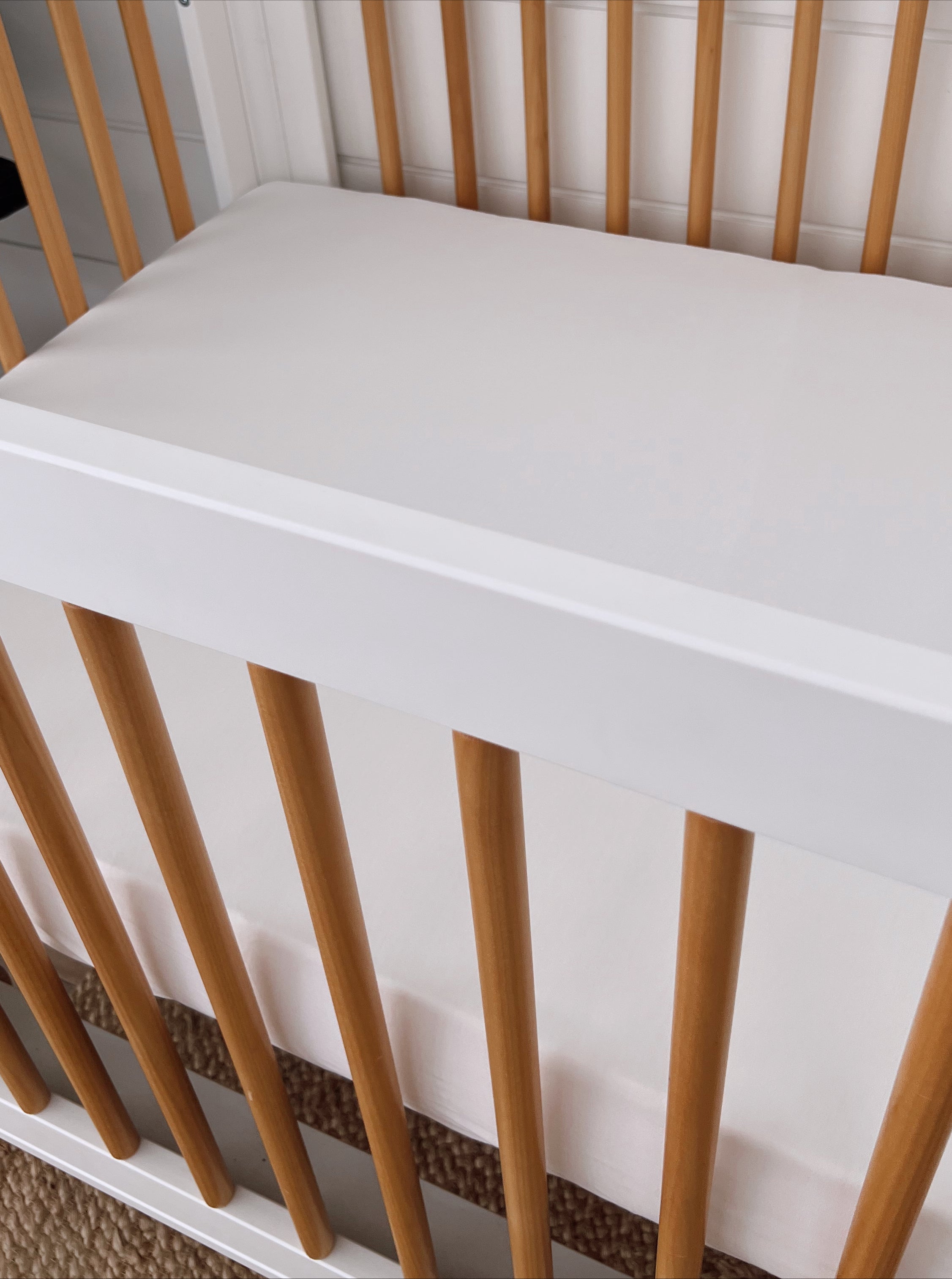 White crib with wooden slats and a plain mattress, set against a light background. Ideal for a nursery.