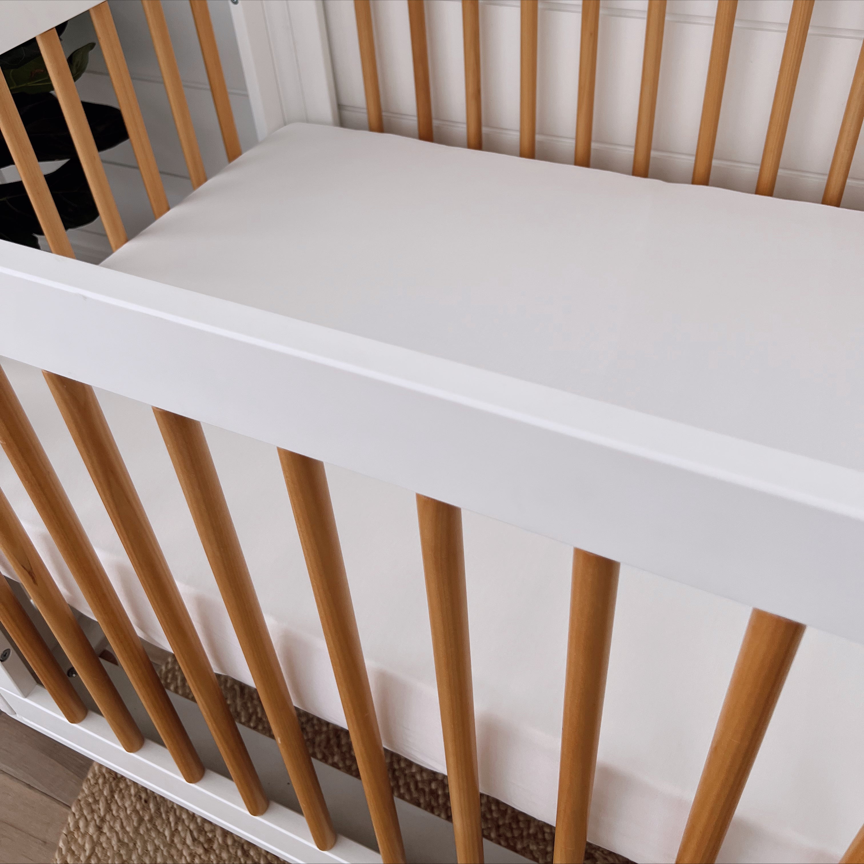 White crib with wooden slats and a plain mattress, set against a light background. Ideal for a nursery.