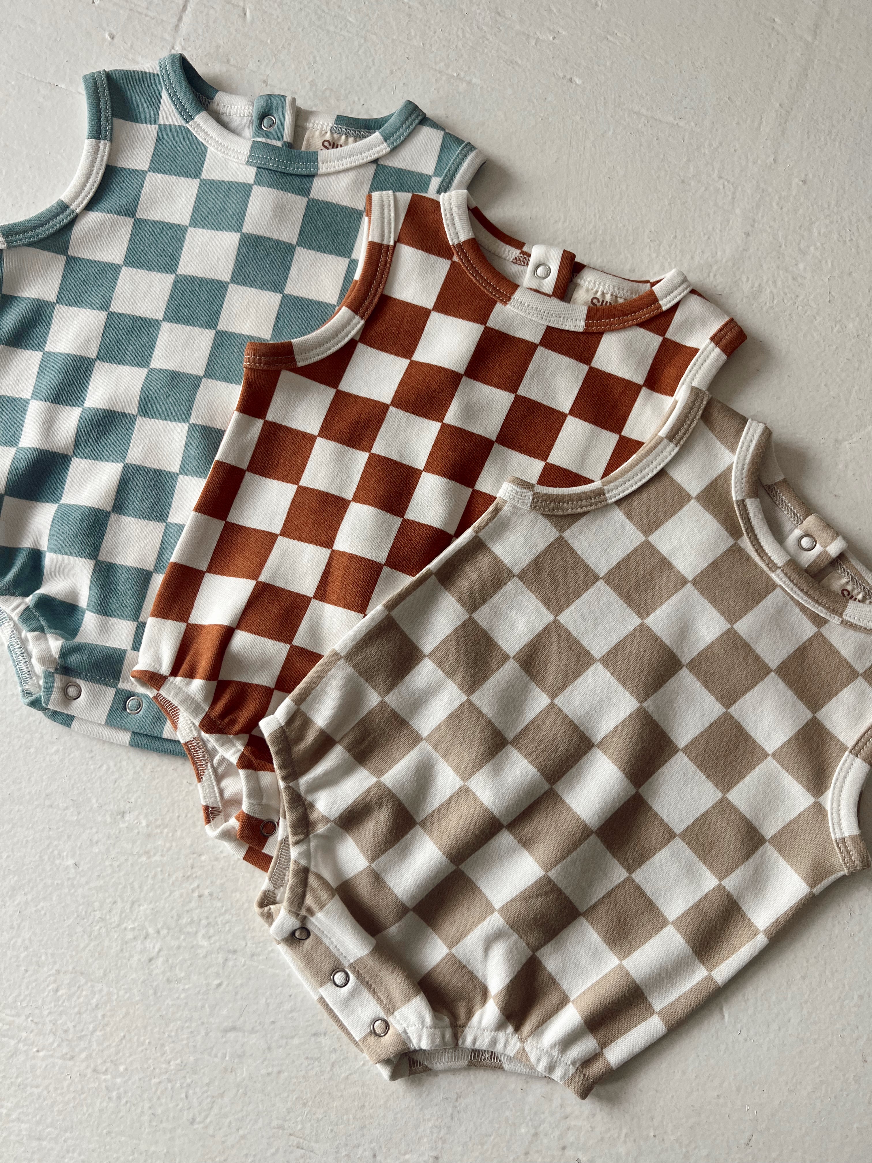 Three checked baby rompers in teal, rust, and beige on a neutral background.