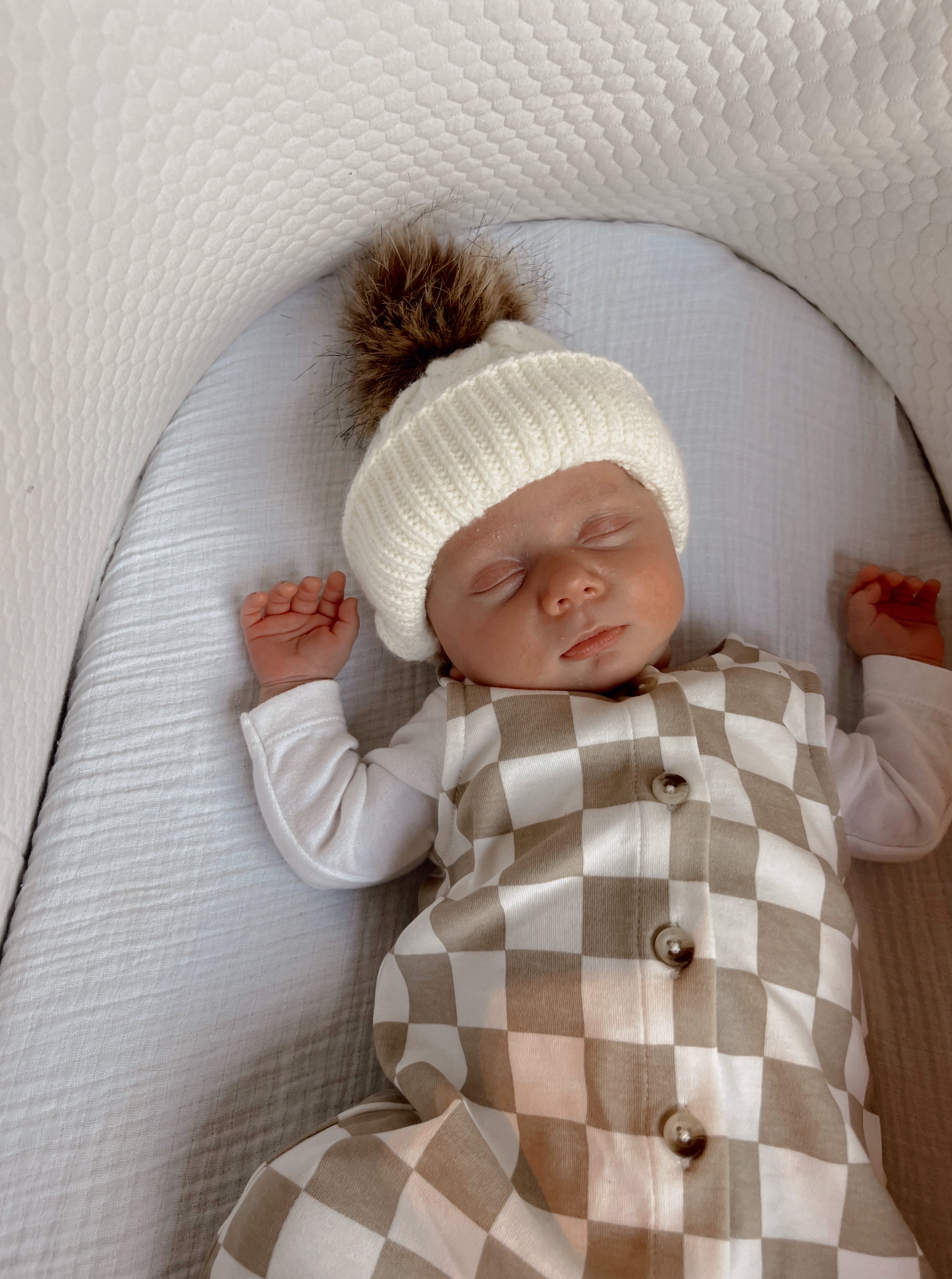 Sleeping baby in a cozy white hat on a textured blanket, dressed in a checkered outfit.