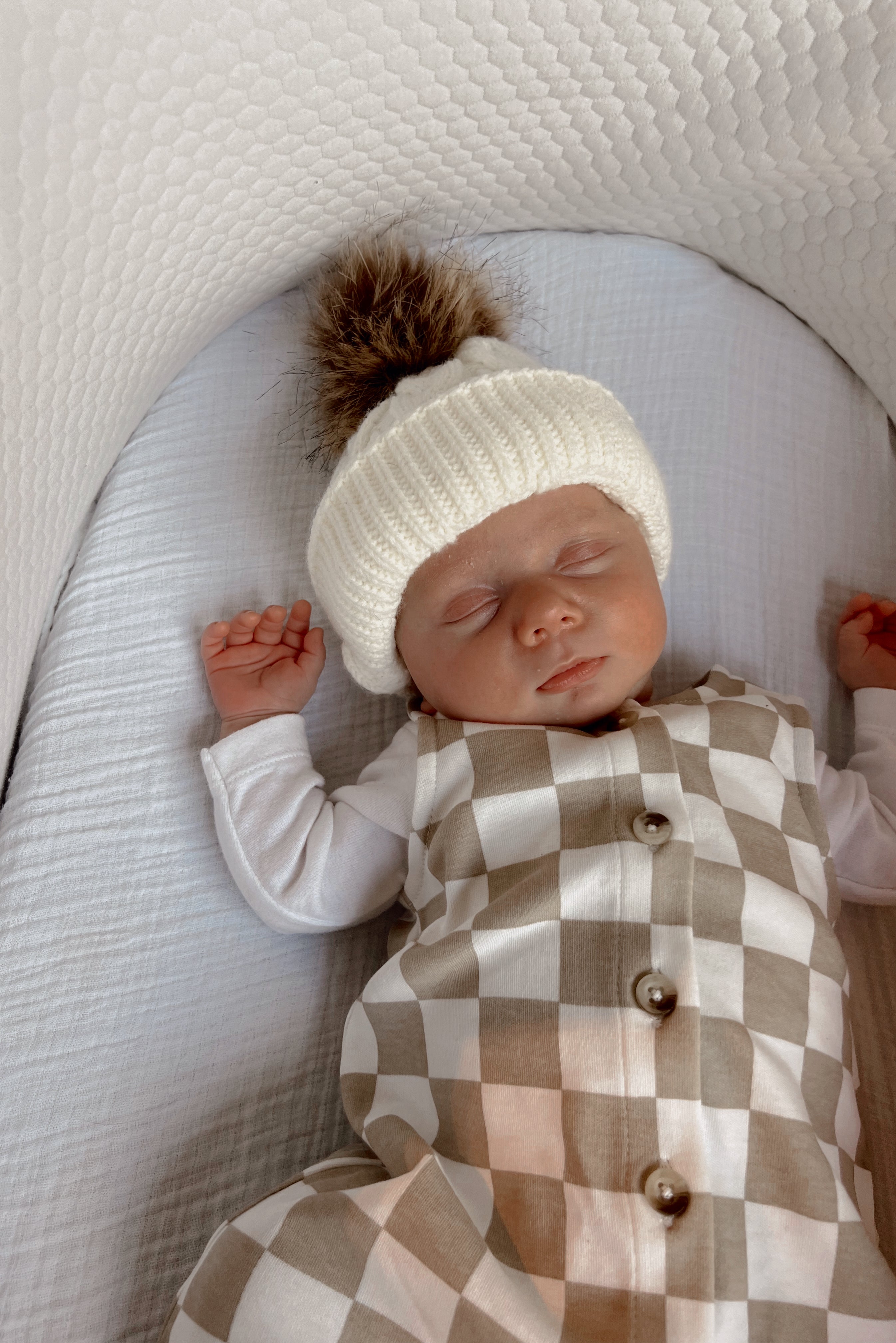 Sleeping baby in a cozy white hat on a textured blanket, dressed in a checkered outfit.