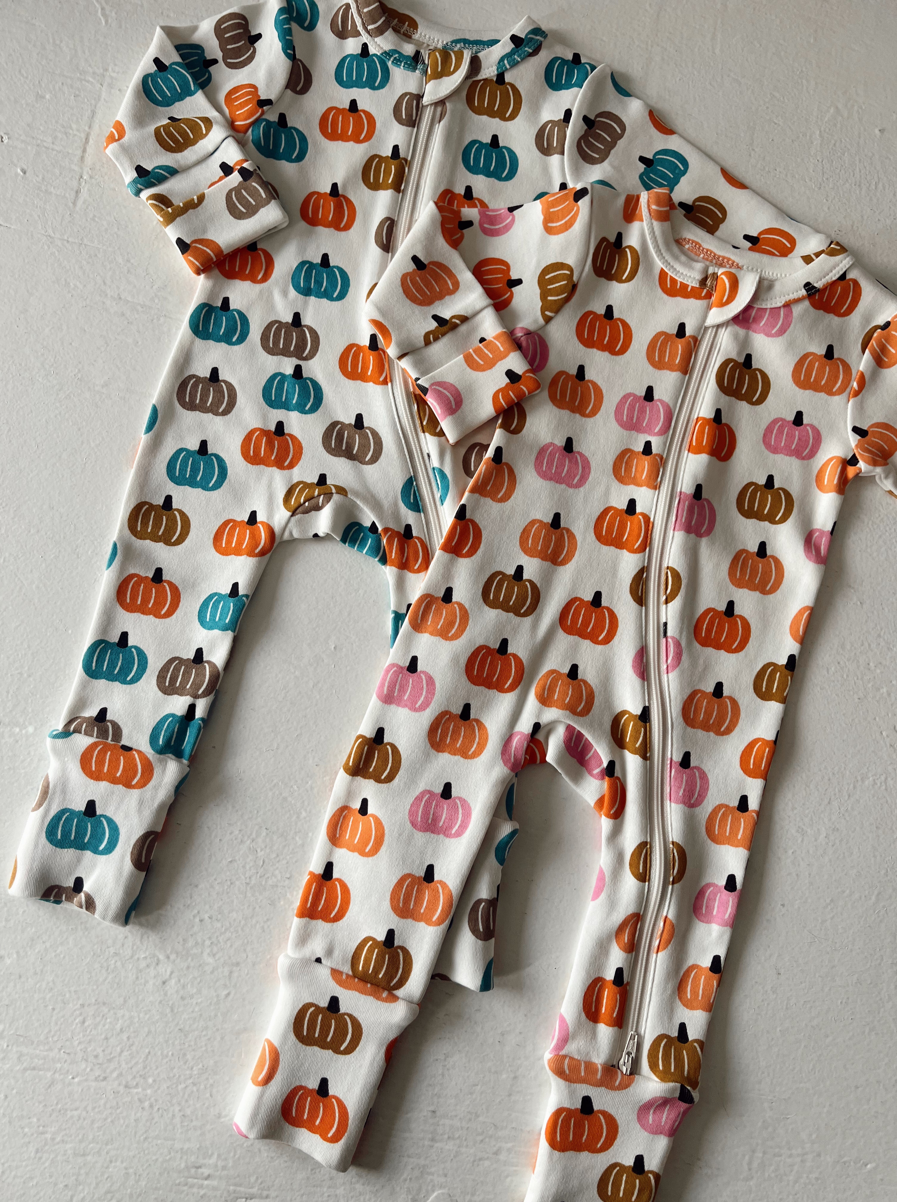 Two infant onesies featuring colorful pumpkin patterns, one with a zip front, laid out on a white surface.