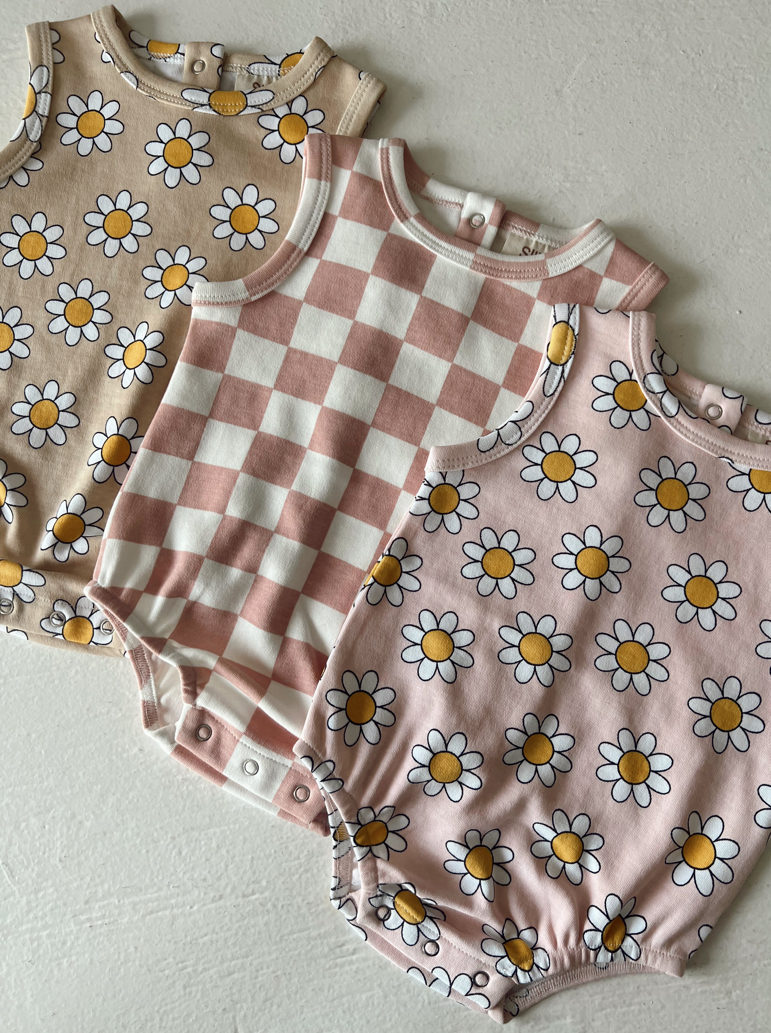 Three baby onesies featuring floral patterns and a checkered design in soft pastel colors.