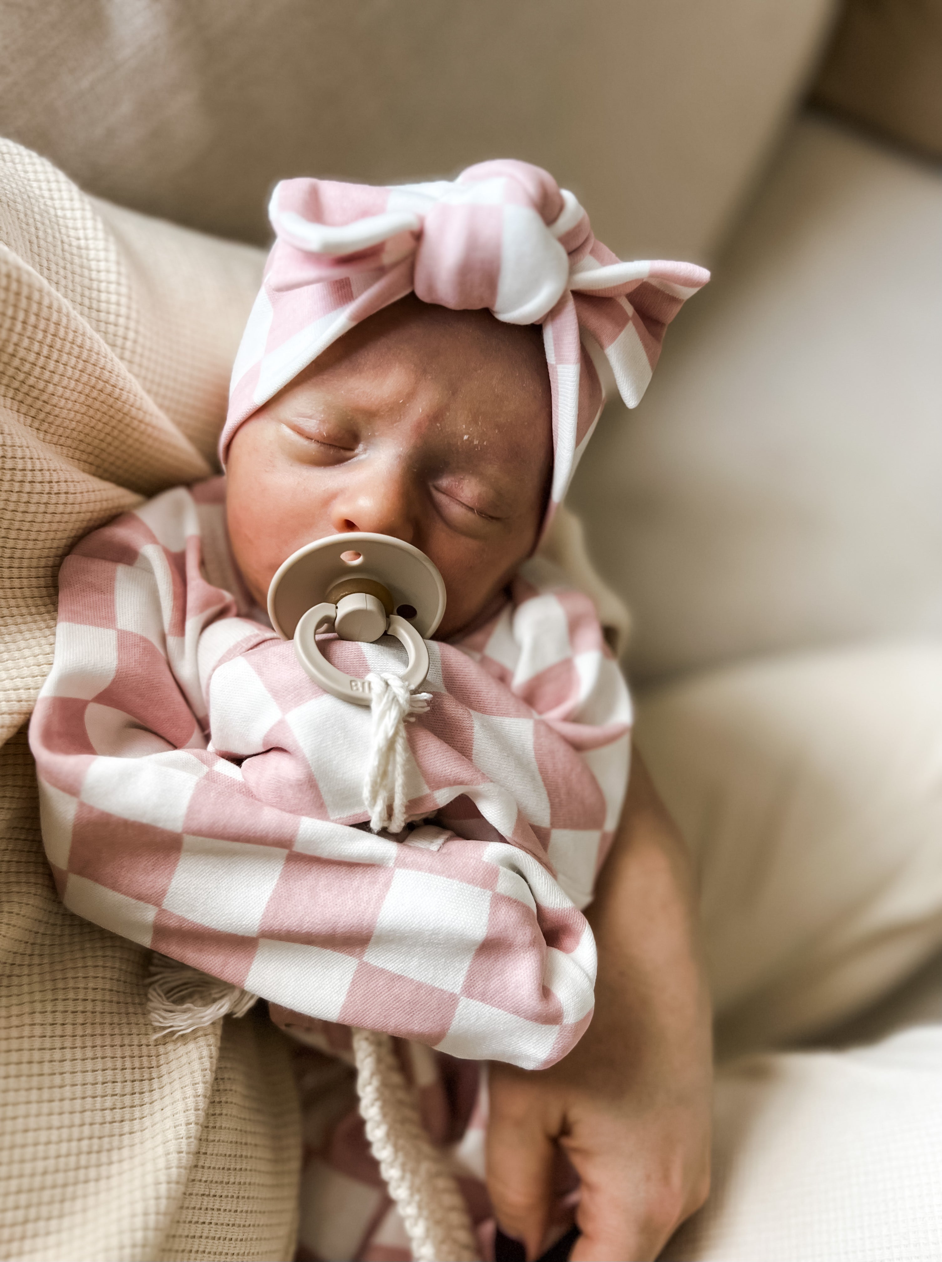 Newborn baby sleeping in a pink checkered outfit with a matching headband and pacifier, held in a caregiver's arms.