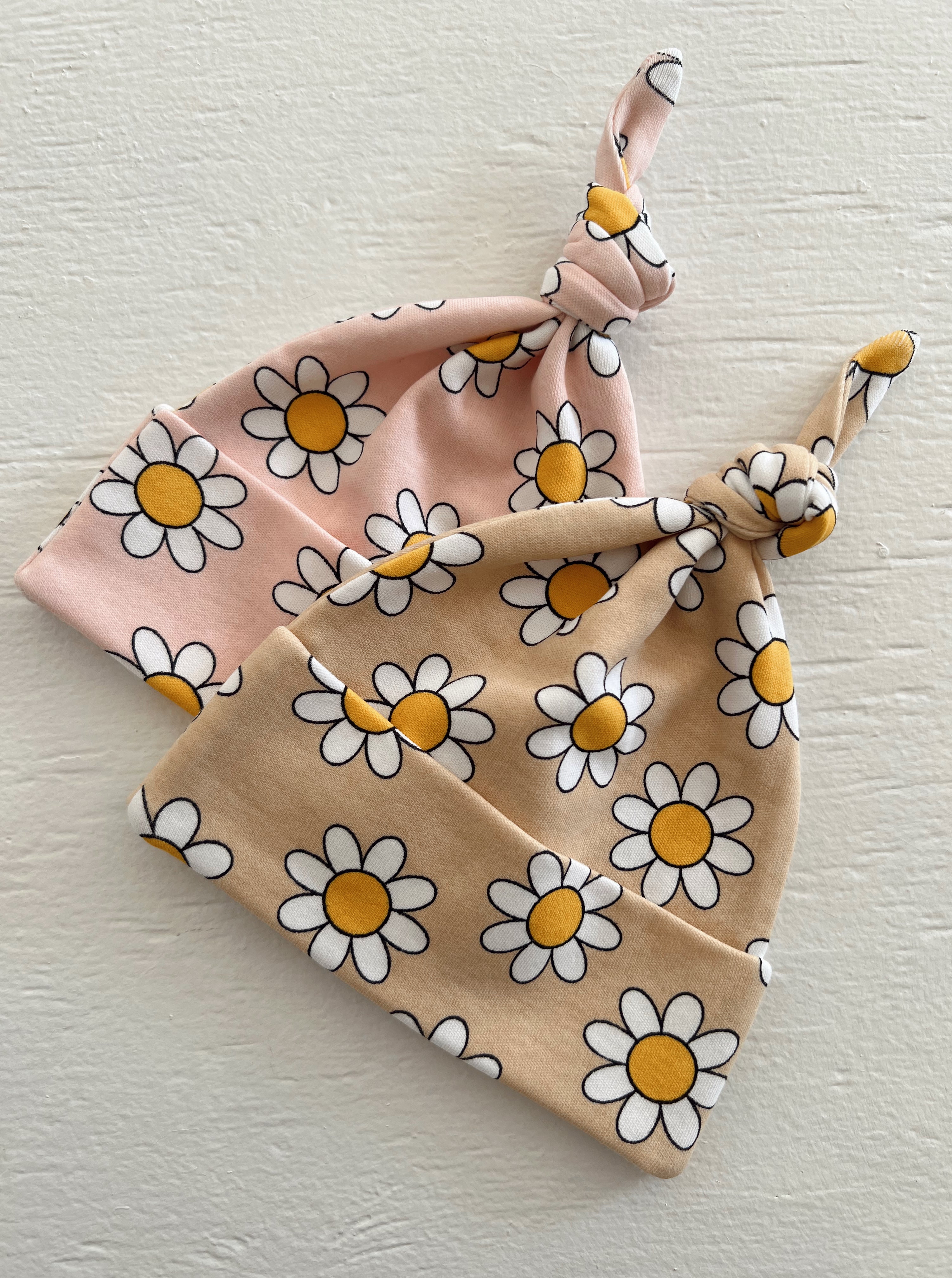 Two flower-patterned baby beanies, one in pink and one in beige, tied into knots.