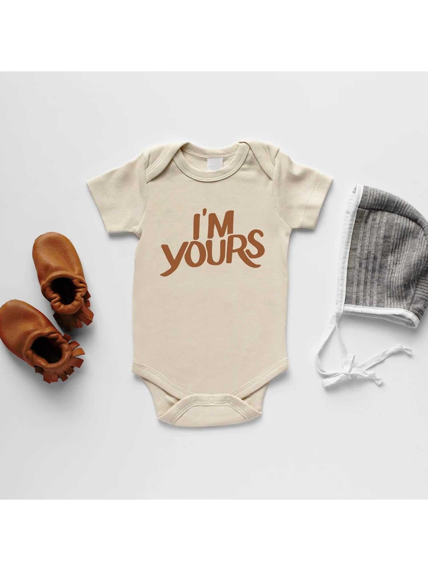 Beige baby onesie with "I'm Yours" print, leather booties, and a gray knit hat on a white background.