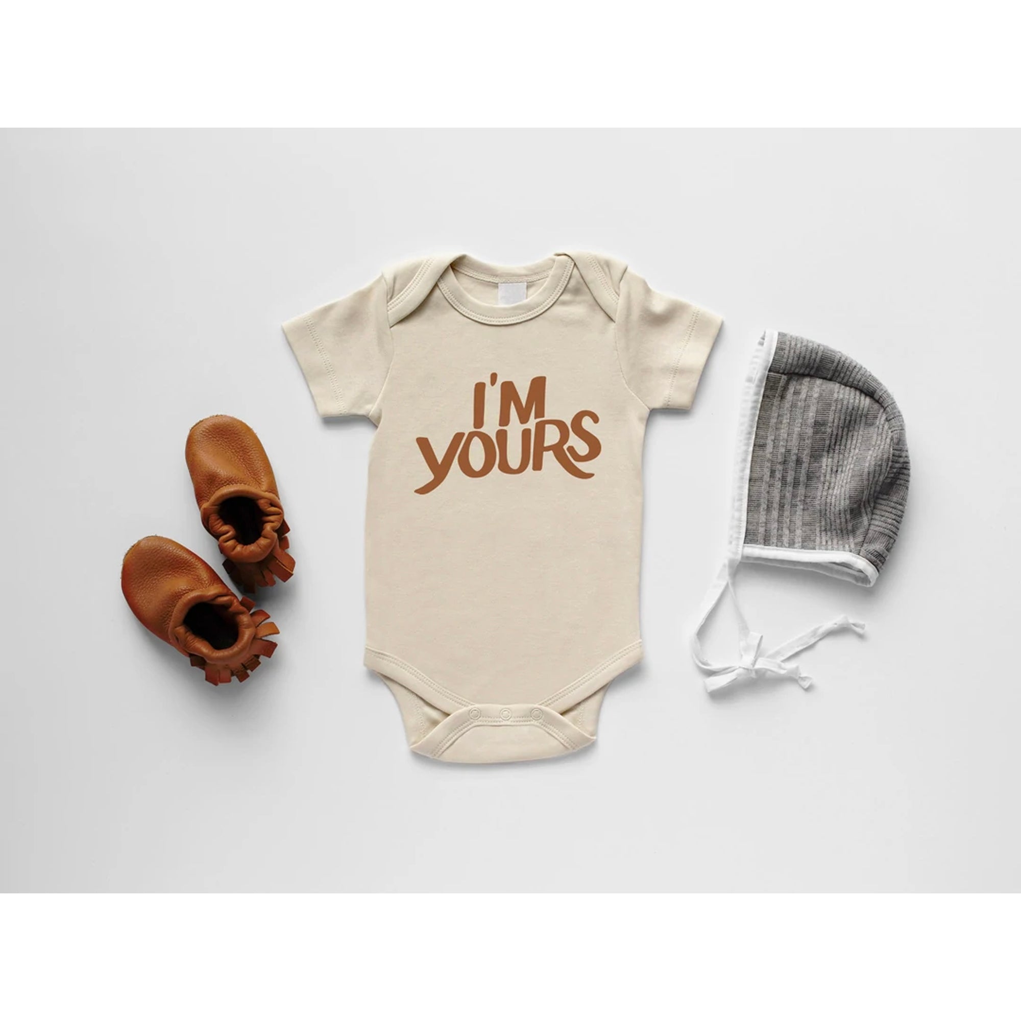 Beige baby onesie with "I'm Yours" print, leather booties, and a gray knit hat on a white background.