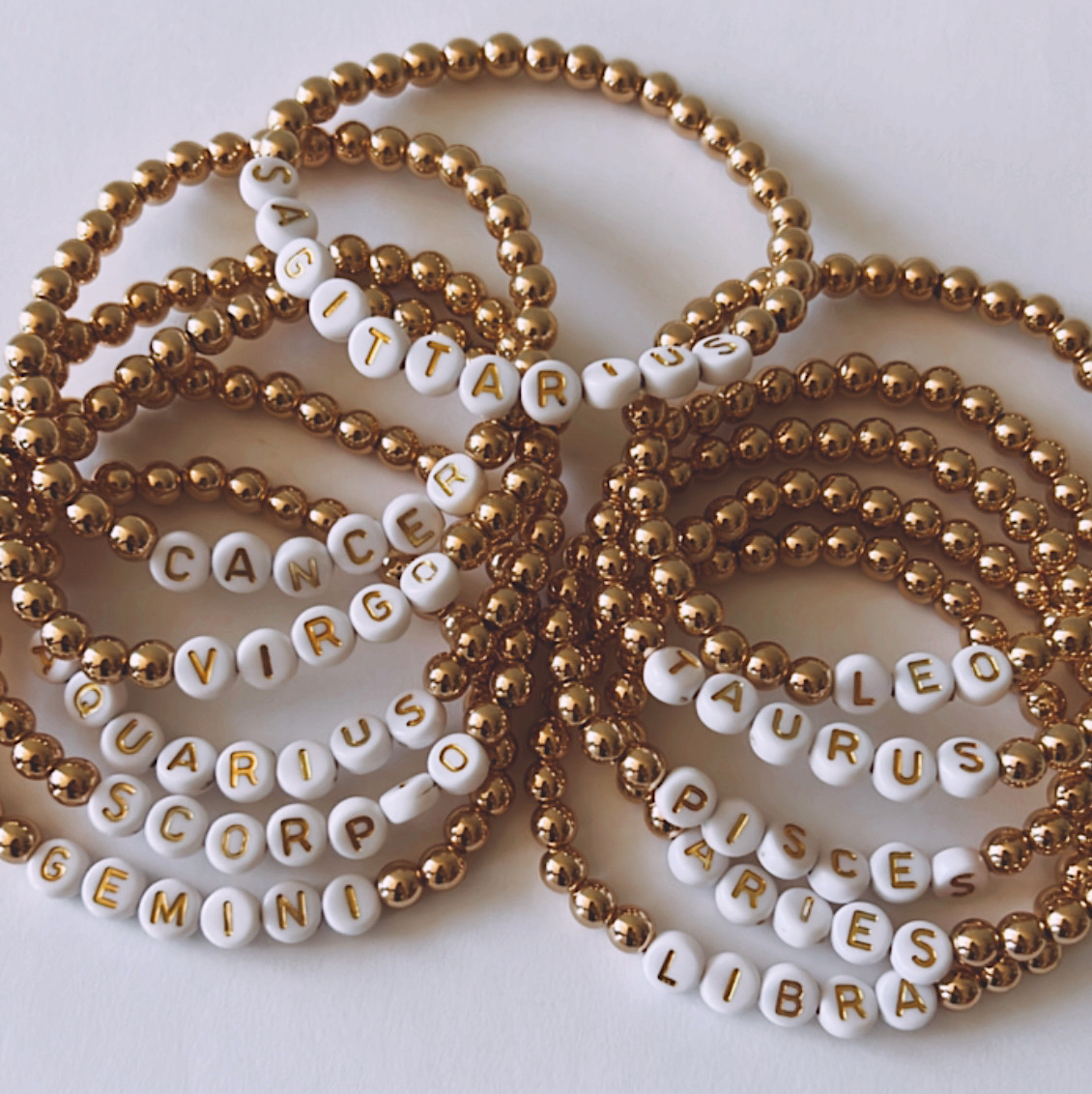 Gold bead bracelets with white letter beads displaying zodiac signs.