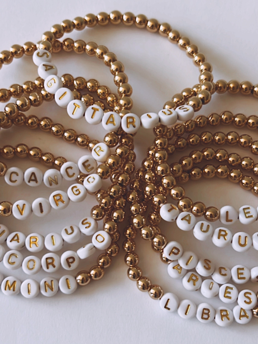 Assorted handmade bracelets featuring zodiac signs in white letters on gold beads.