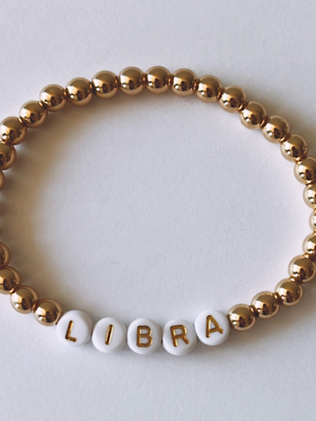 Gold and white beaded bracelet featuring the word “LIBRA” spelled out in letter beads.