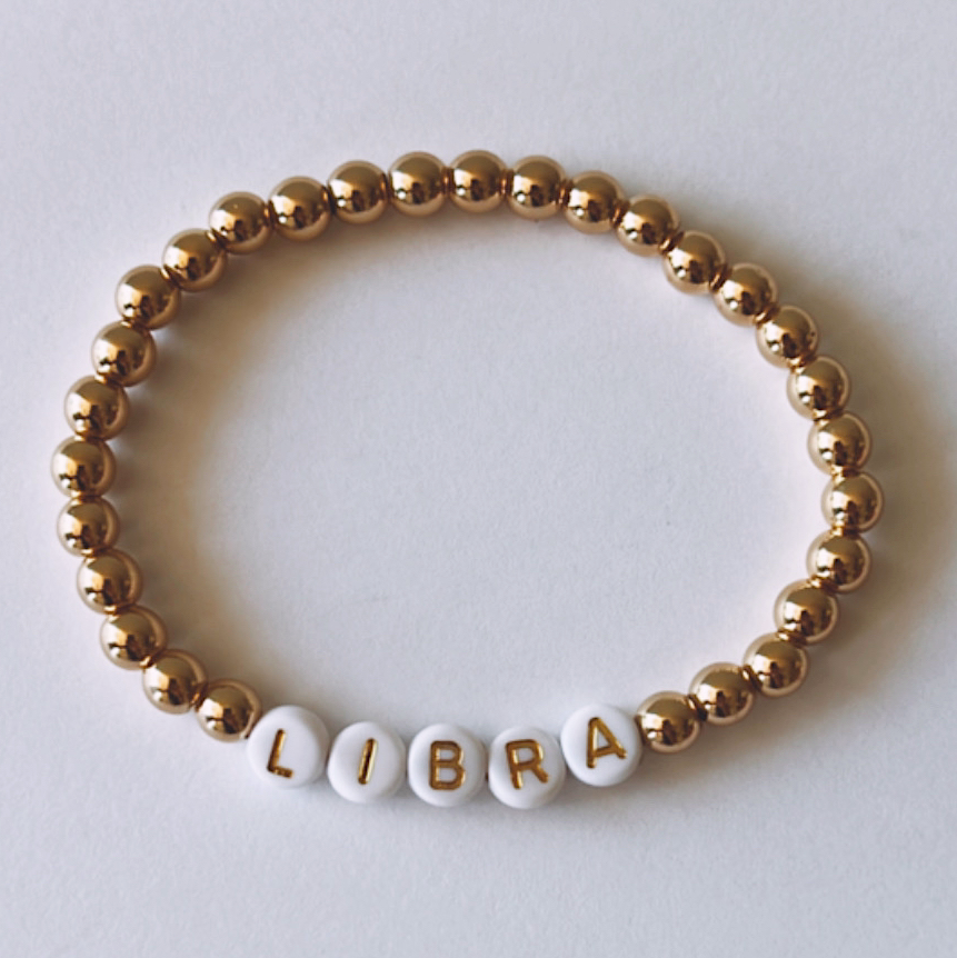 Gold and white beaded bracelet featuring the word “LIBRA” spelled out in letter beads.