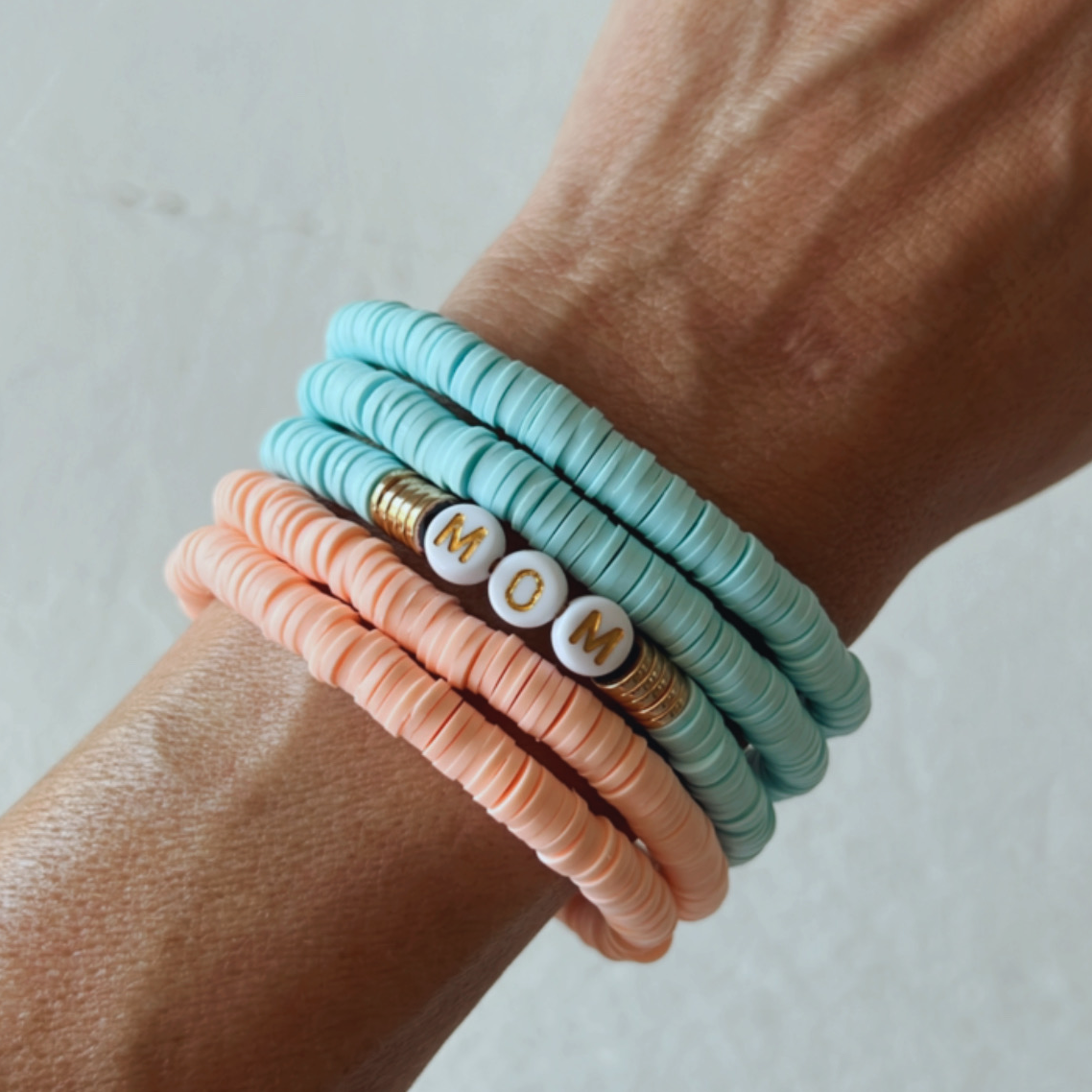 Colorful stacked bracelets featuring a charm that spells "MOM" on a wrist.
