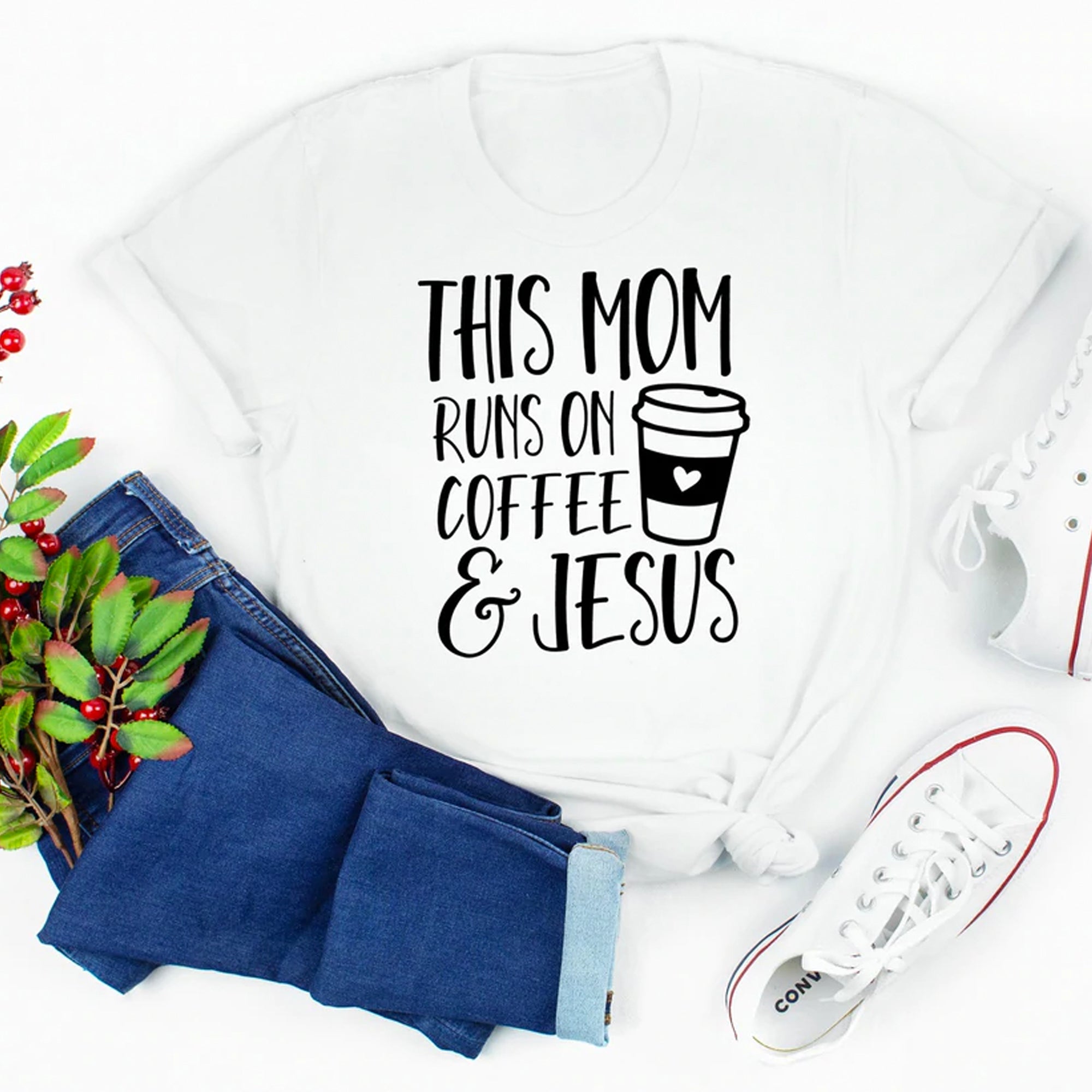White t-shirt with "This Mom Runs on Coffee & Jesus" graphic, styled with jeans and sneakers, surrounded by green leaves.