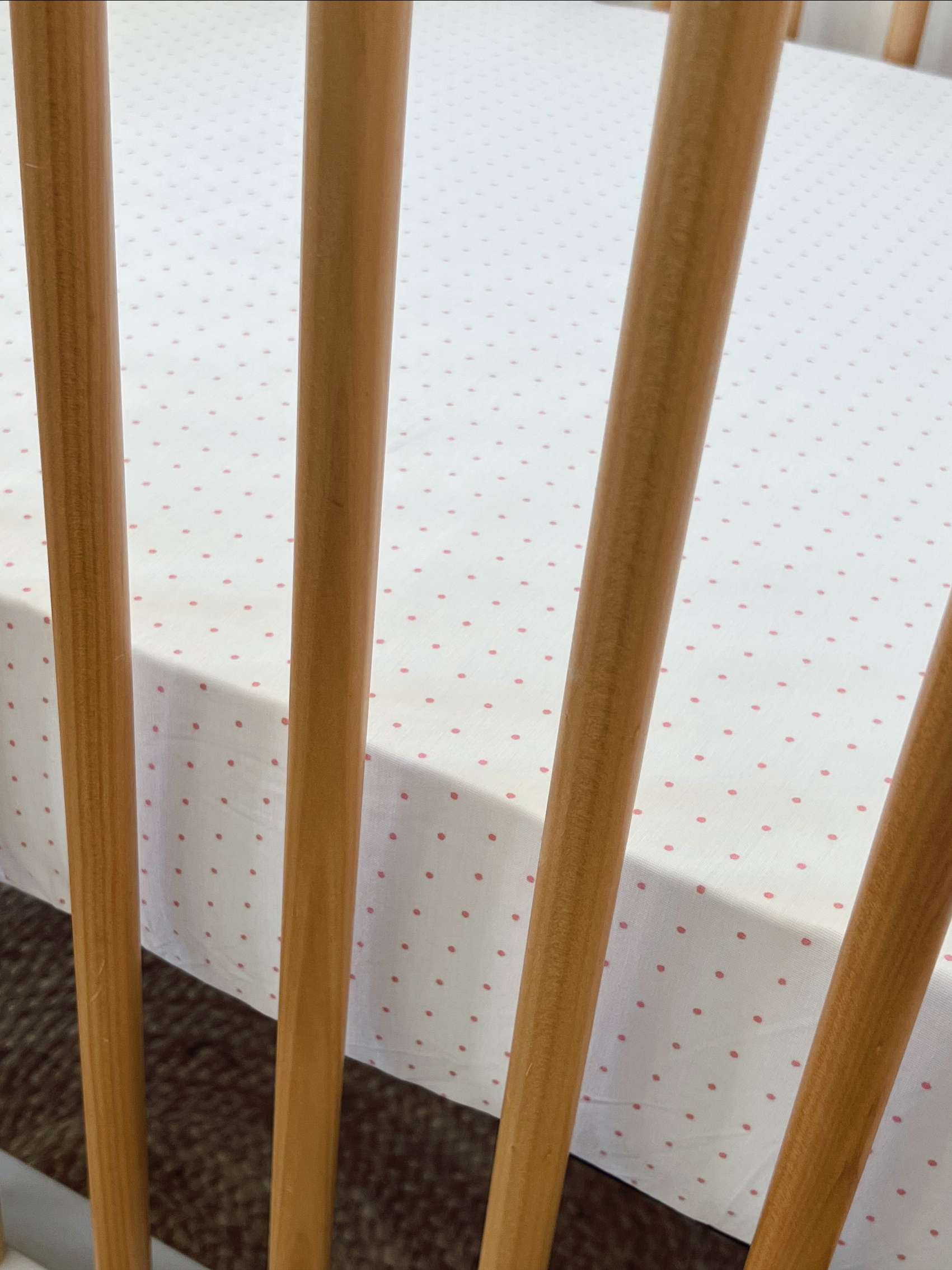 Crib with soft white bedding featuring small pink polka dots, seen through wooden slats.