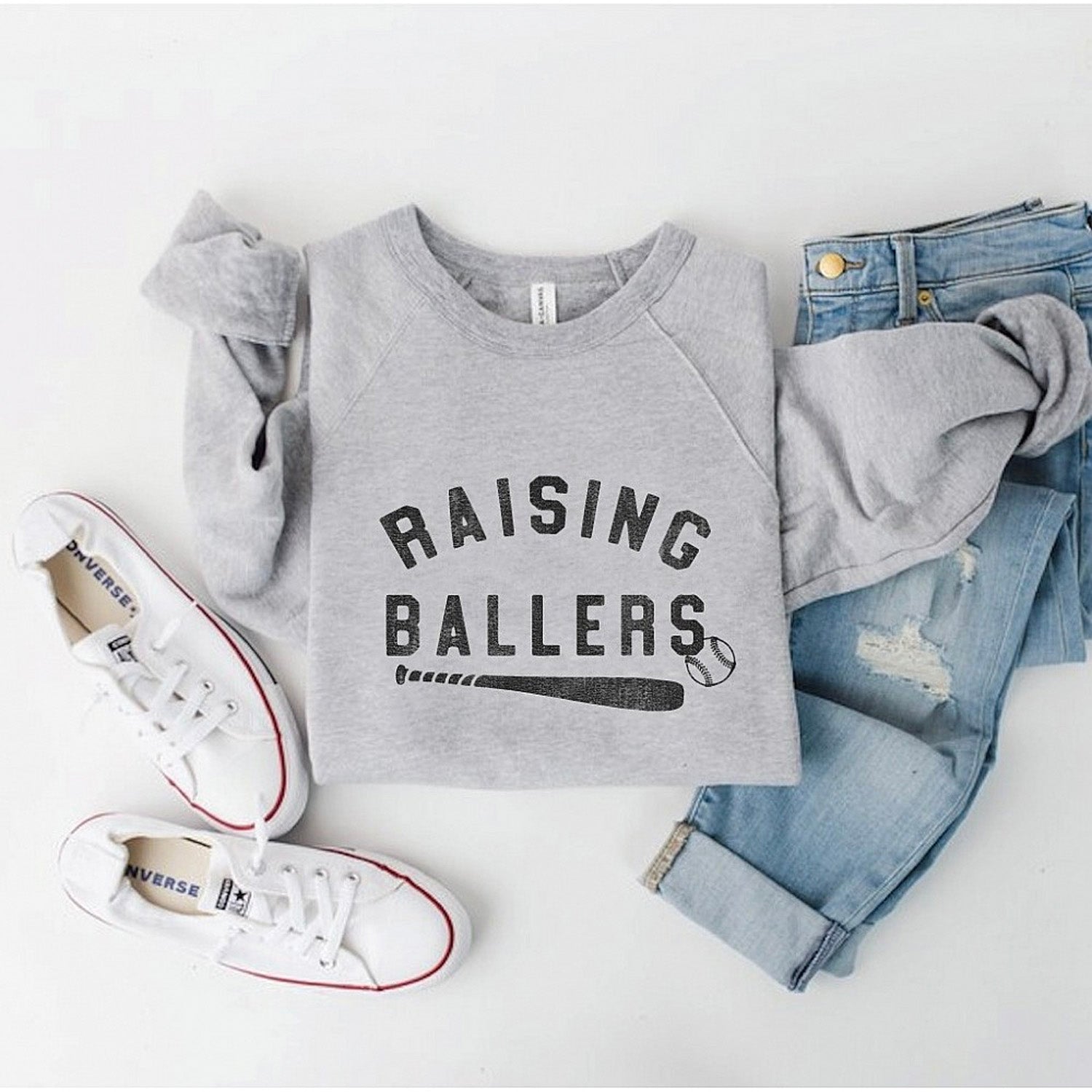 Gray sweatshirt with "Raising Ballers" text, paired with jeans and white sneakers on a neutral background.