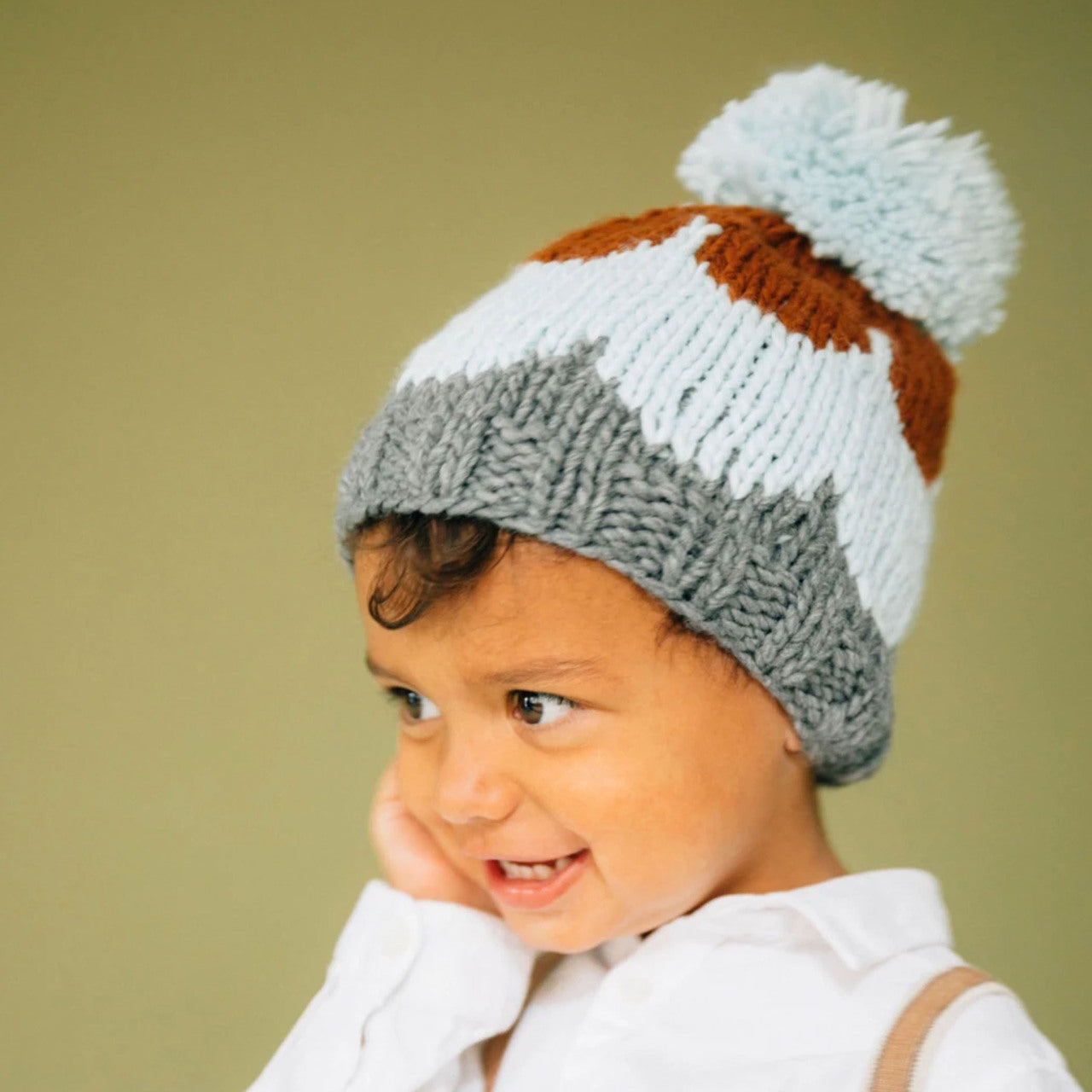 Smiling child wearing a colorful knitted hat with a pom-pom against a soft green background.