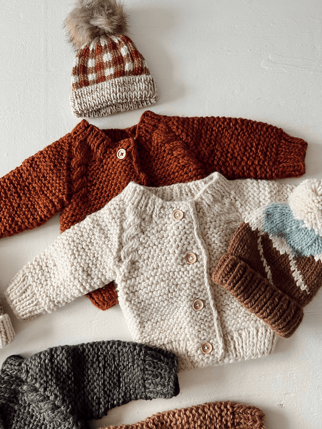 Knitted baby sweaters in various colors with a checked hat, arranged on a light surface.