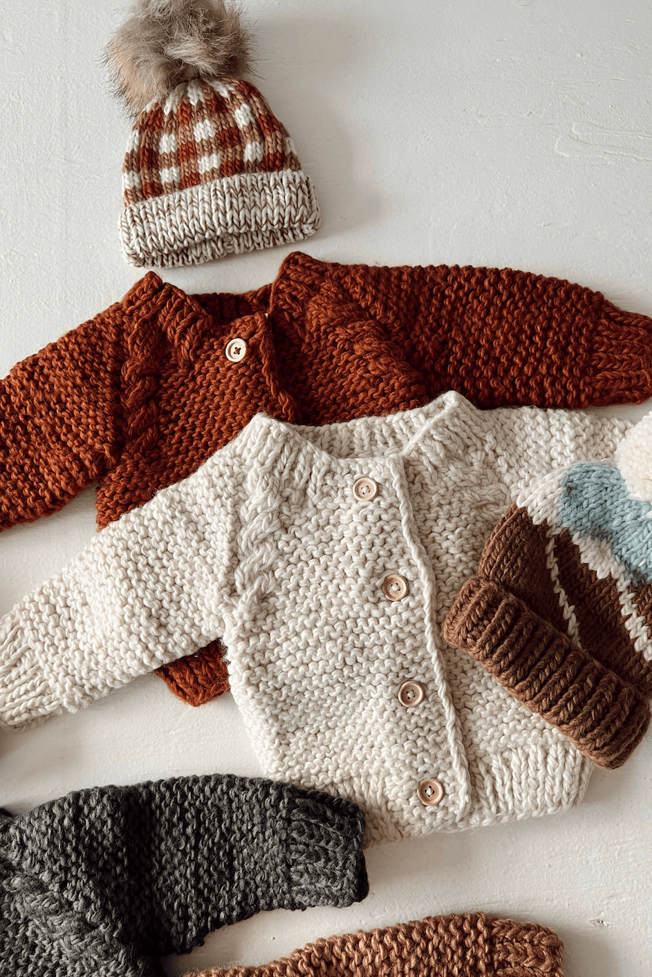 Knitted baby sweaters in various colors with a checked hat, arranged on a light surface.
