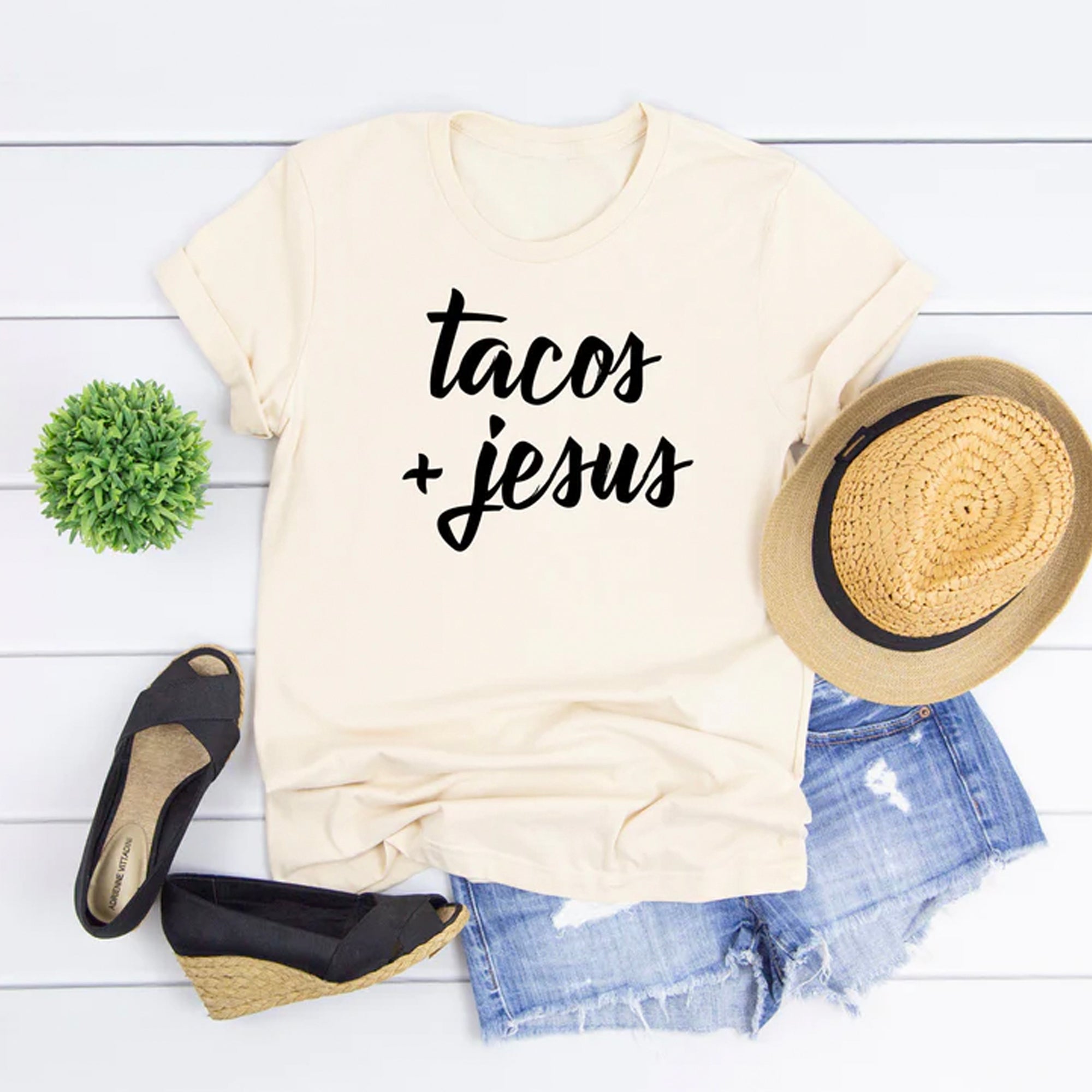 Beige t-shirt with "tacos + Jesus" print, paired with denim shorts, black wedge sandals, and a straw hat.