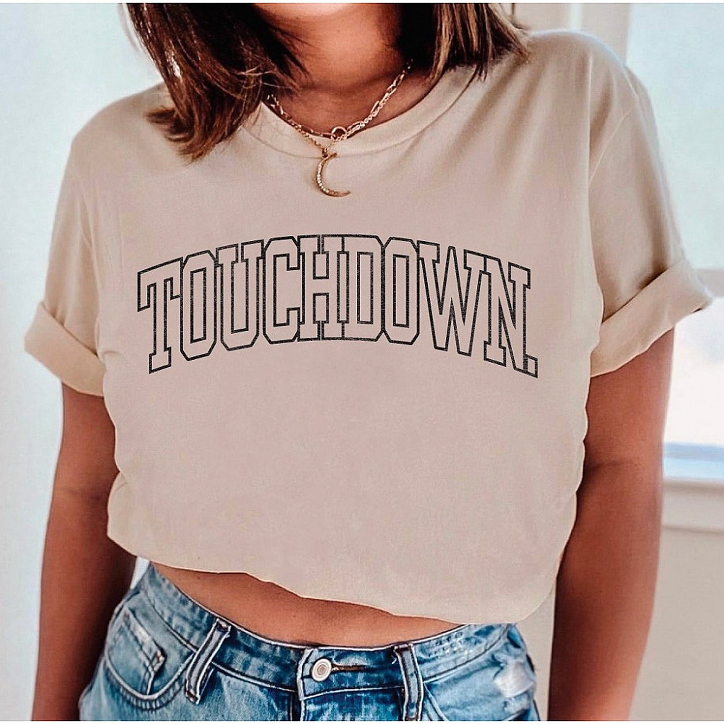 Woman wearing a cropped cream t-shirt with the word "TOUCHDOWN" in bold letters, paired with denim shorts.