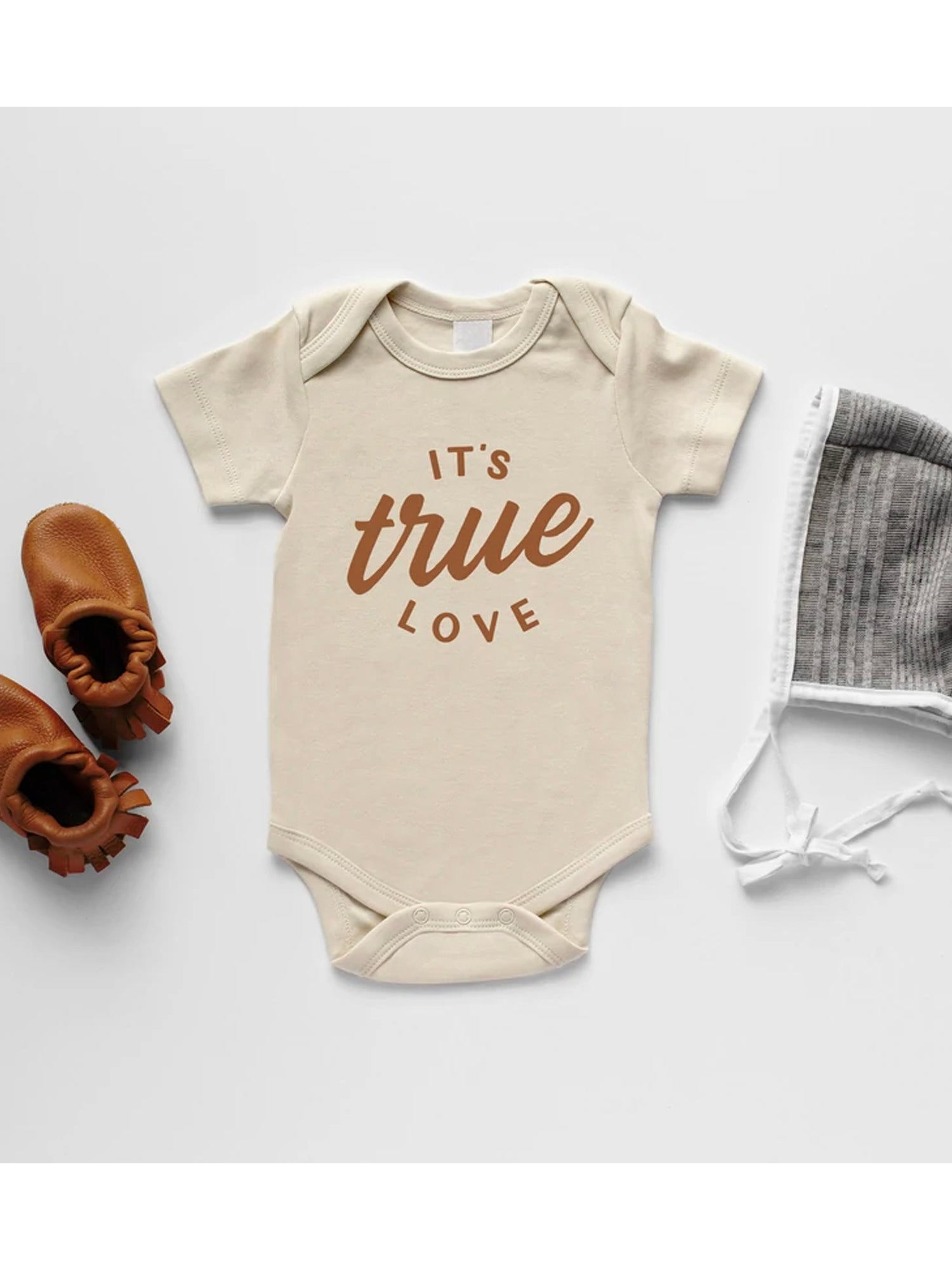 Beige baby onesie with "It's true love" text, paired with brown baby shoes and a gray bonnet, against a white background.
