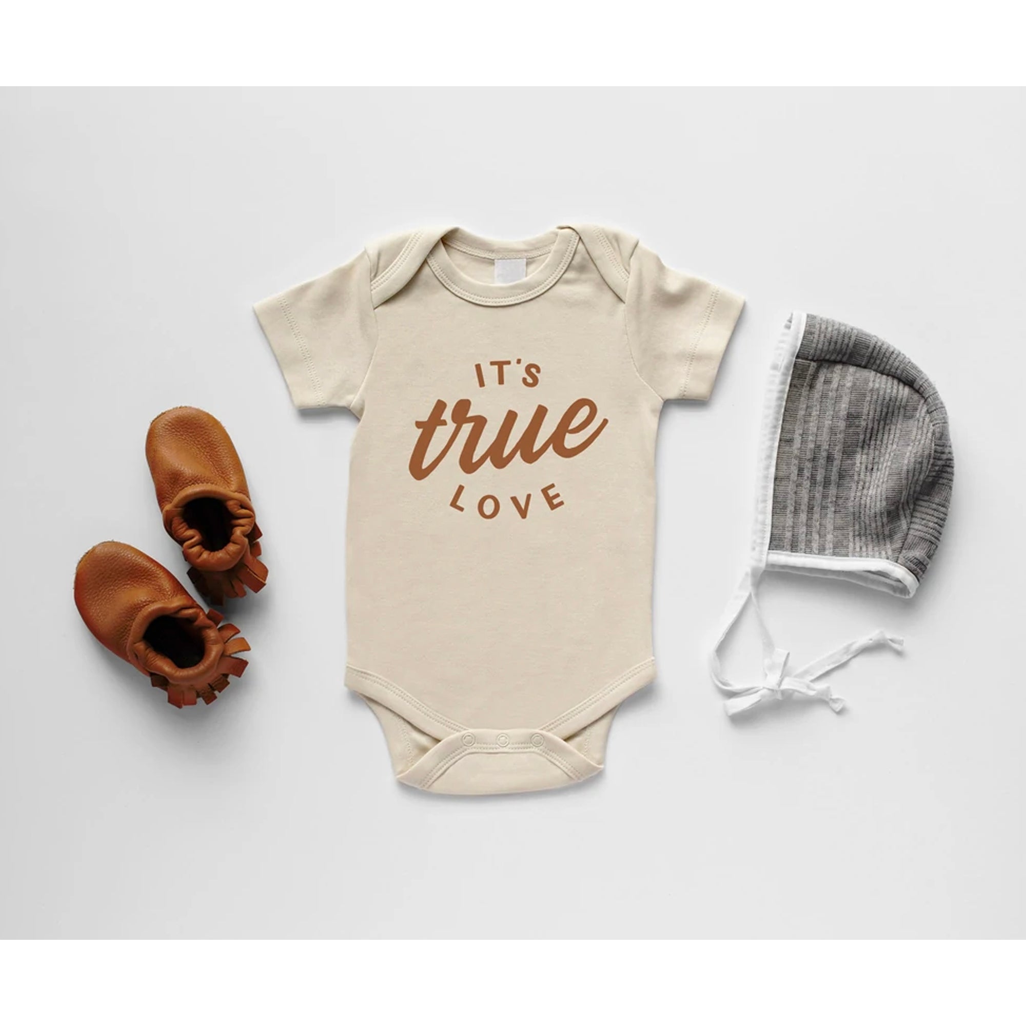 Beige baby onesie with "It's true love" text, paired with brown baby shoes and a gray bonnet, against a white background.