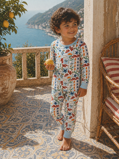 Child in colorful pajamas holds a lemon, smiling on a sunlit terrace overlooking the sea.