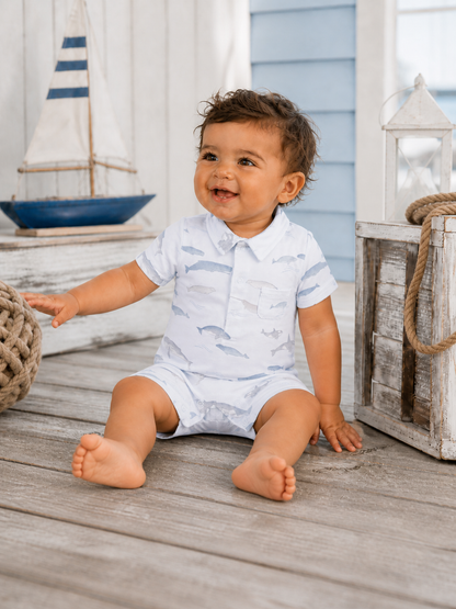 Smiling toddler in a fish-patterned outfit sitting on wooden flooring near nautical decor.