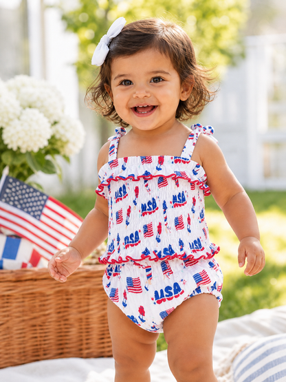 Smiling toddler in patriotic swimsuit with USA print, standing outdoors near flags and flowers.