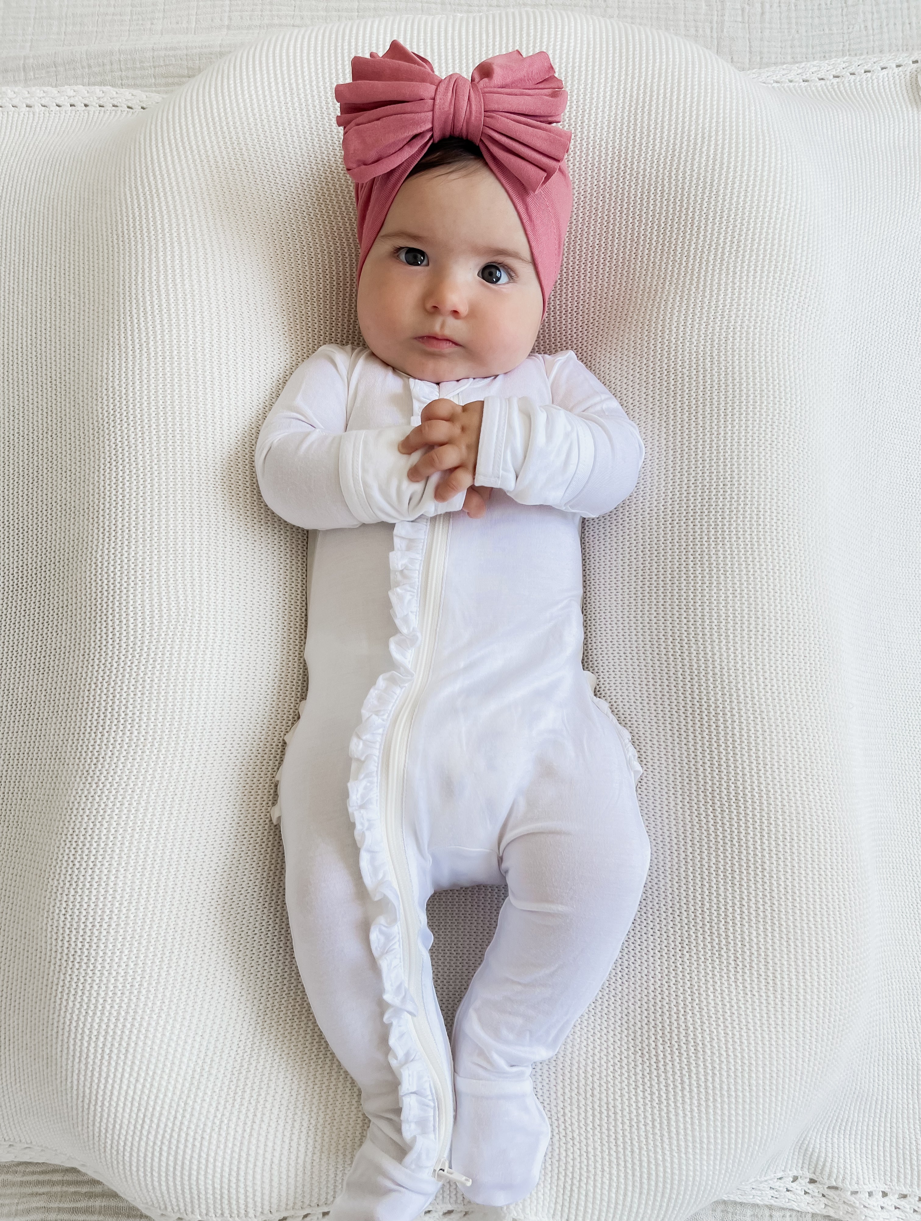 Baby in white onesie with ruffles, wearing a pink bow headband, resting on a textured white surface.