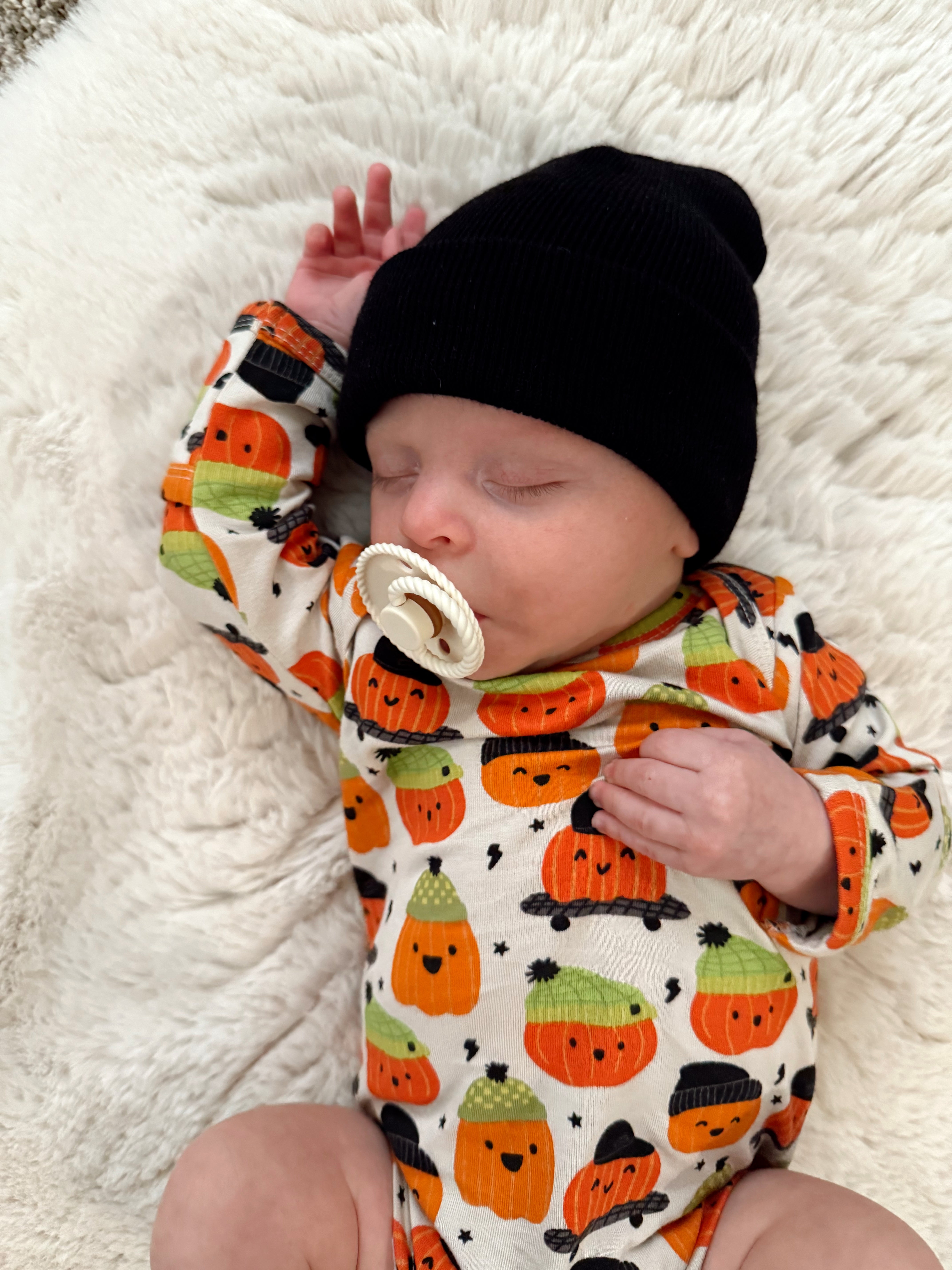 Baby asleep on a soft blanket, wearing a black beanie and an outfit with colorful pumpkin patterns.