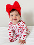 Smiling baby in a red bow headband and matching outfit with a white background.