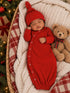 Sleeping baby in red outfit with hat, snuggled in a cozy blanket next to a teddy bear and holiday decor.