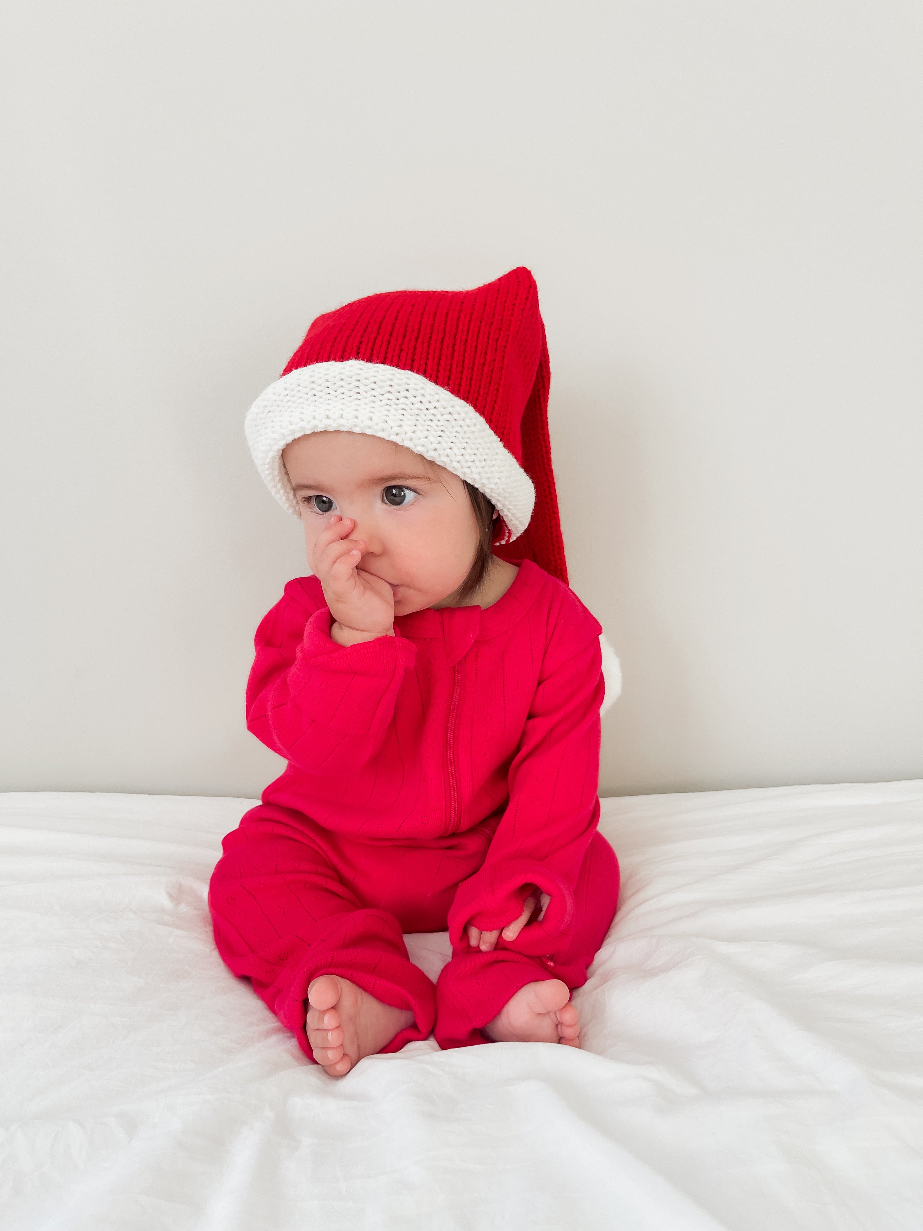 Baby in a red outfit with a Santa hat, sitting on a white blanket and sucking thumb. Cheerful holiday vibe.