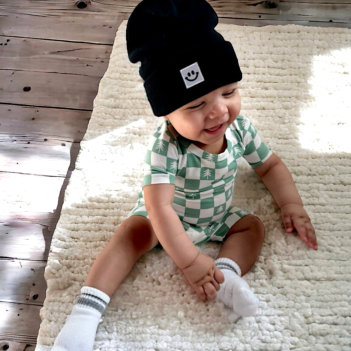 Smiling baby in a patterned outfit and beanie, sitting on a textured rug with soft lighting.
