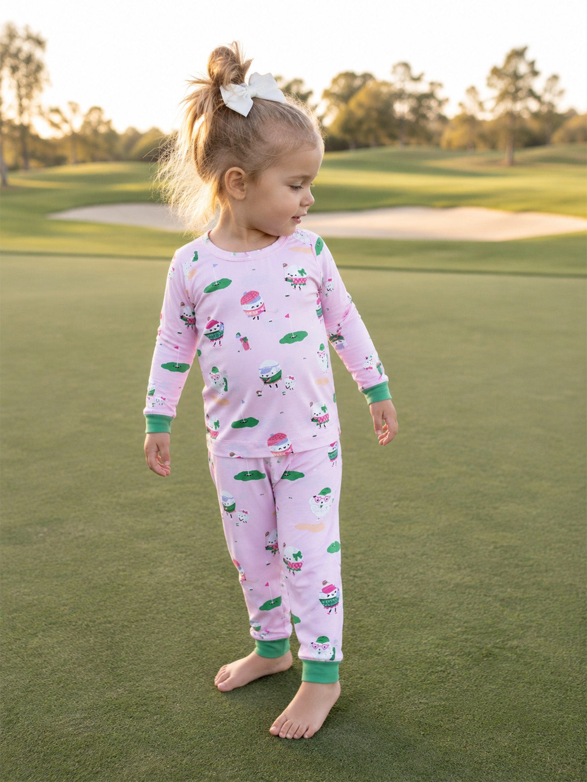 Toddler in pink pajamas with whimsical patterns stands barefoot on a grassy field, smiling at the camera.
