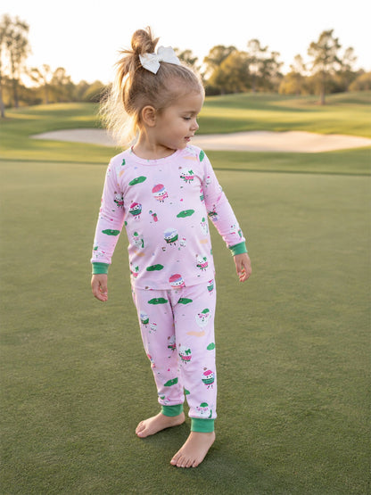 Toddler in pink pajamas with whimsical patterns stands barefoot on a grassy field, smiling at the camera.
