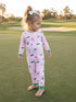 Toddler in pink pajamas with whimsical patterns stands barefoot on a grassy field, smiling at the camera.