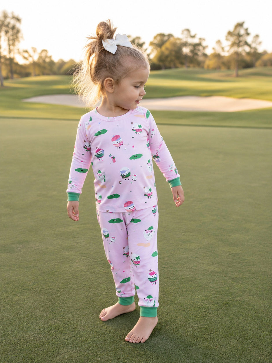 Toddler in pink pajamas with whimsical patterns stands barefoot on a grassy field, smiling at the camera.