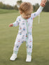 Toddler in playful golf-themed onesie, joyfully walking on a green, held by an adult's hand.